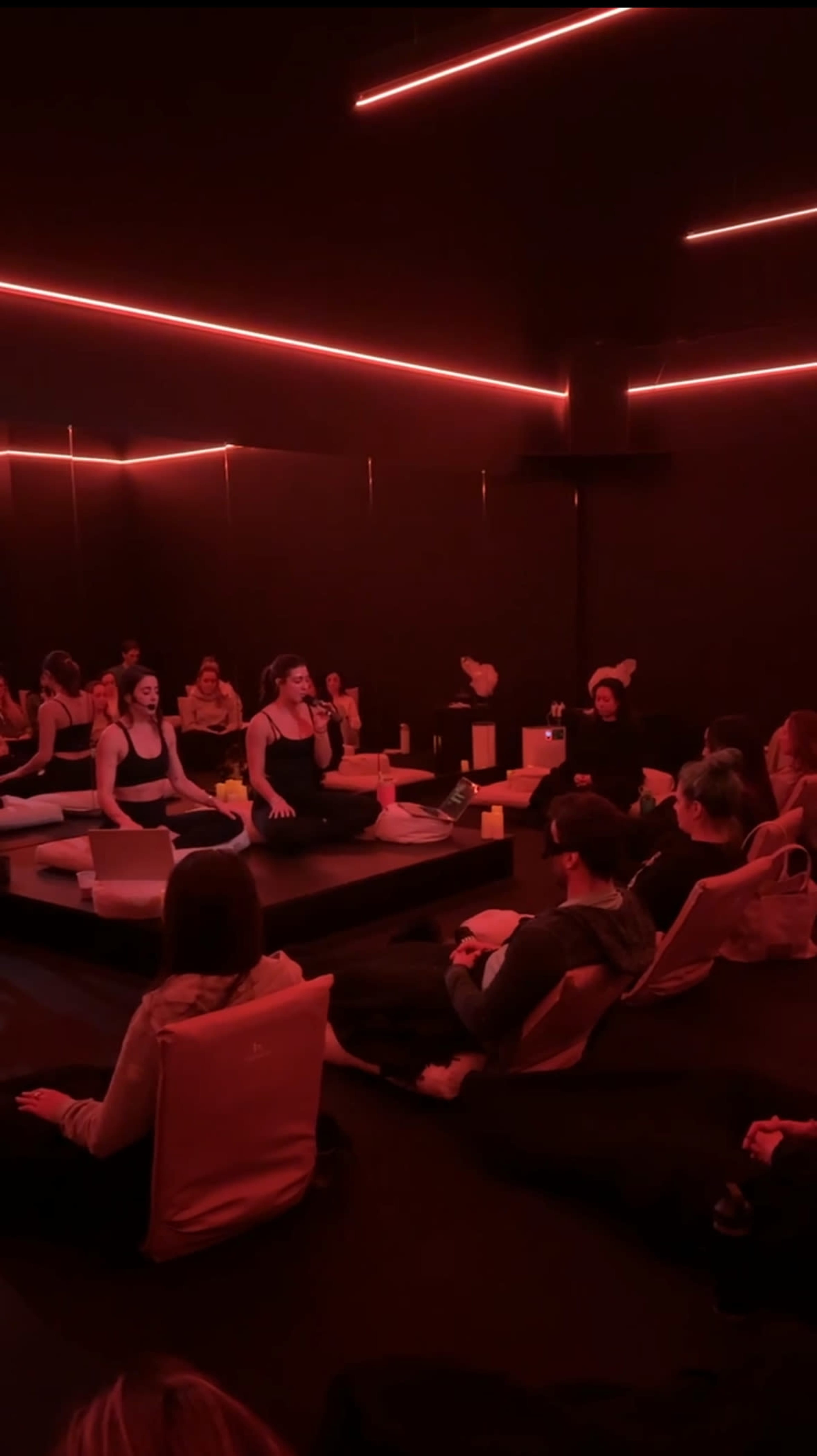 A group of individuals is seated in a dimly lit room with red lighting, engaging in a guided session led by an instructor.