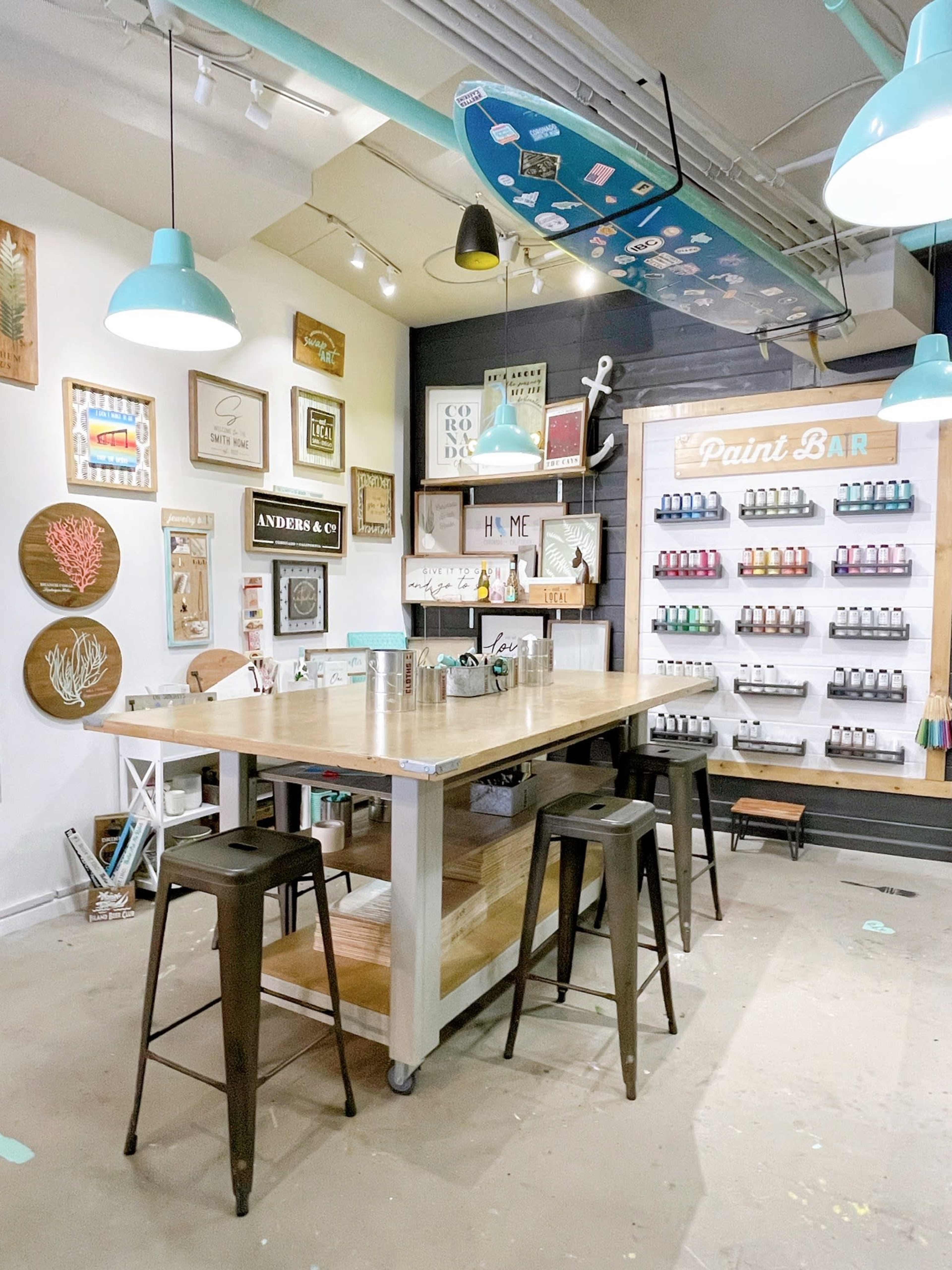 A crafting space featuring a central wooden table surrounded by black stools, with a wall displaying an array of paint bottles and various framed decorations.