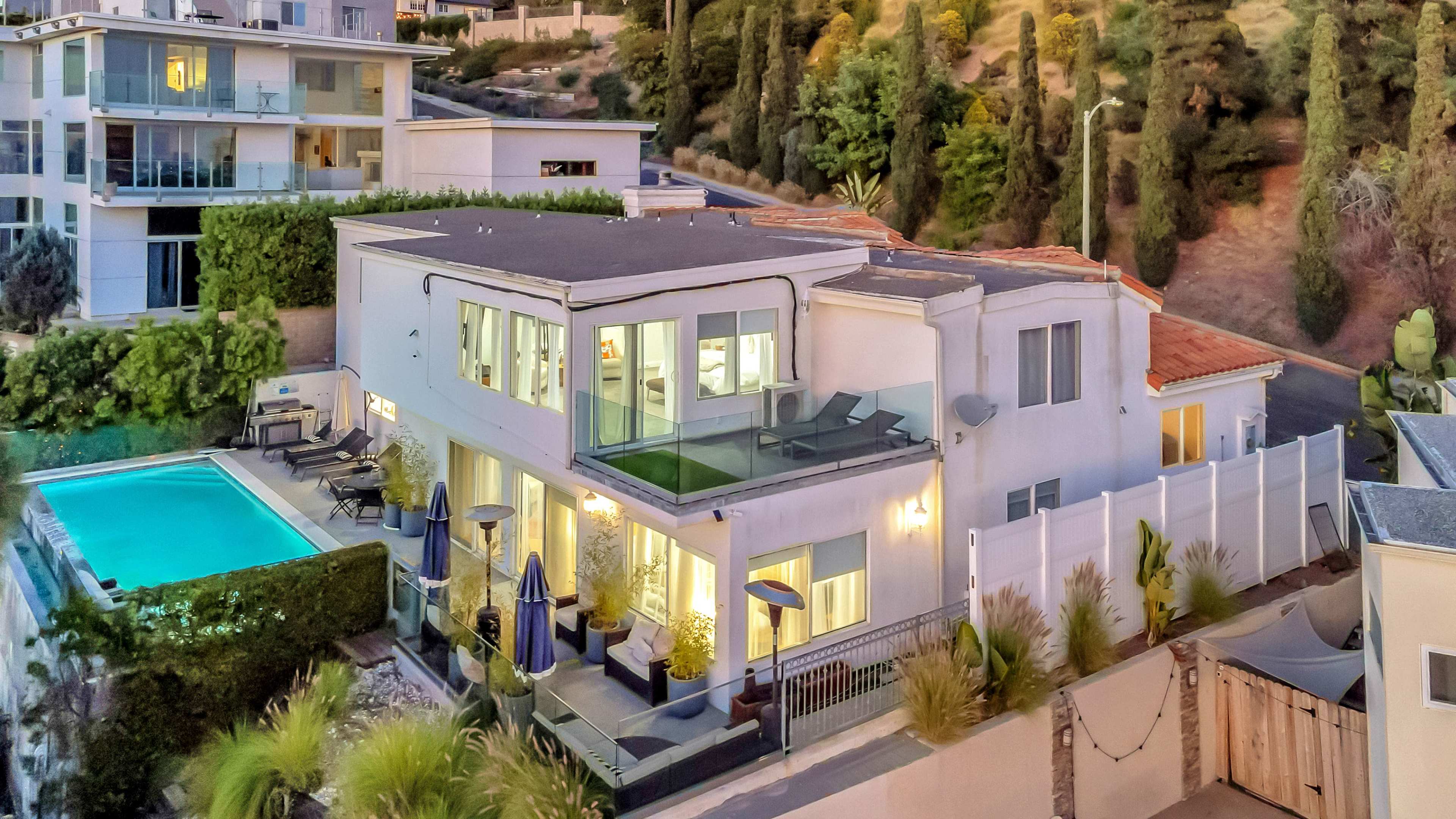 A modern house with large windows overlooks a pool and landscaped yard, situated amidst hilly terrain.