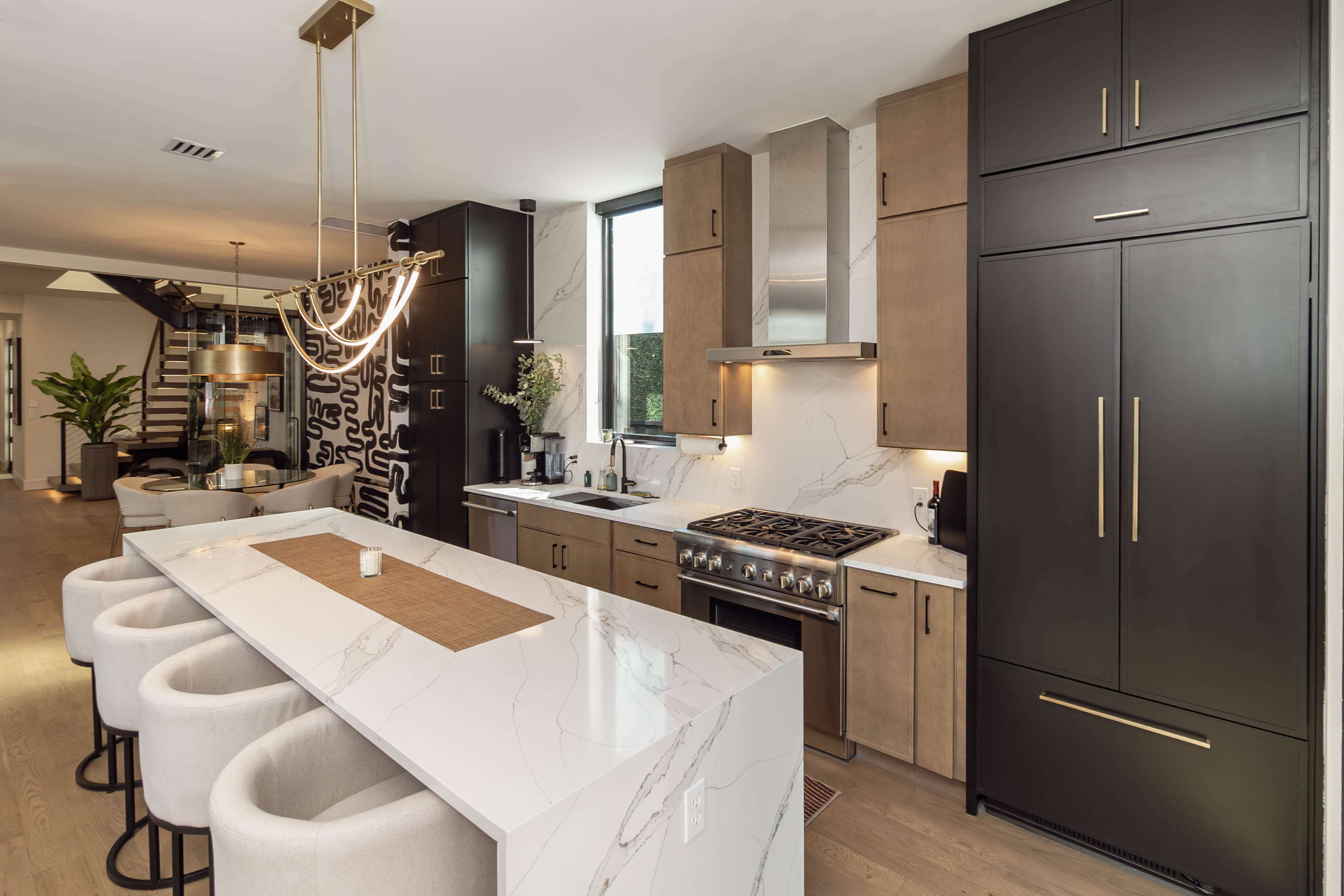 The image shows a modern kitchen featuring a large island with four stools, sleek cabinetry in dark and light tones, and professional-grade appliances.
