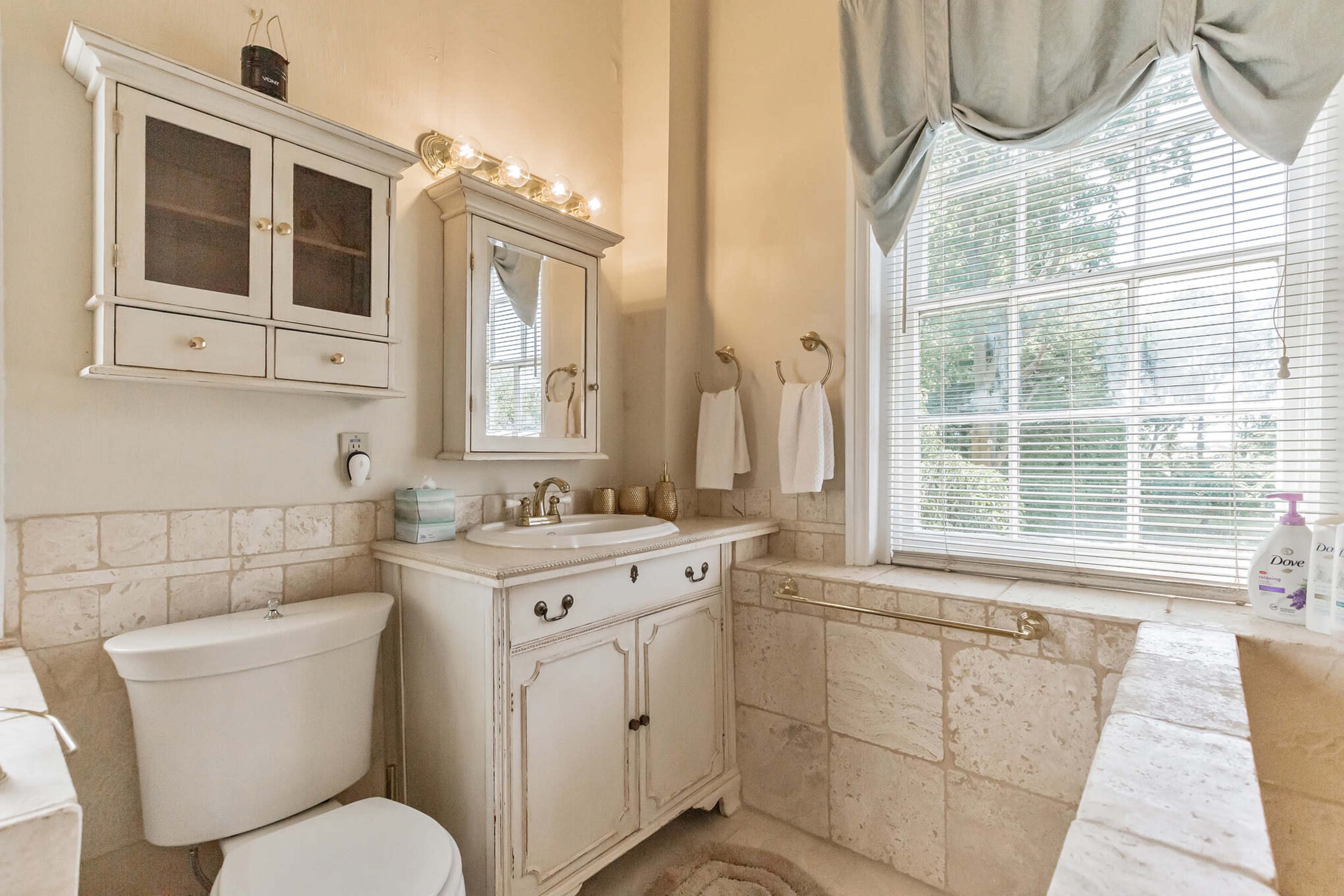 The image shows a bathroom with a white vanity, a toilet, a wall-mounted mirror, and a window covered with a light fabric curtain.