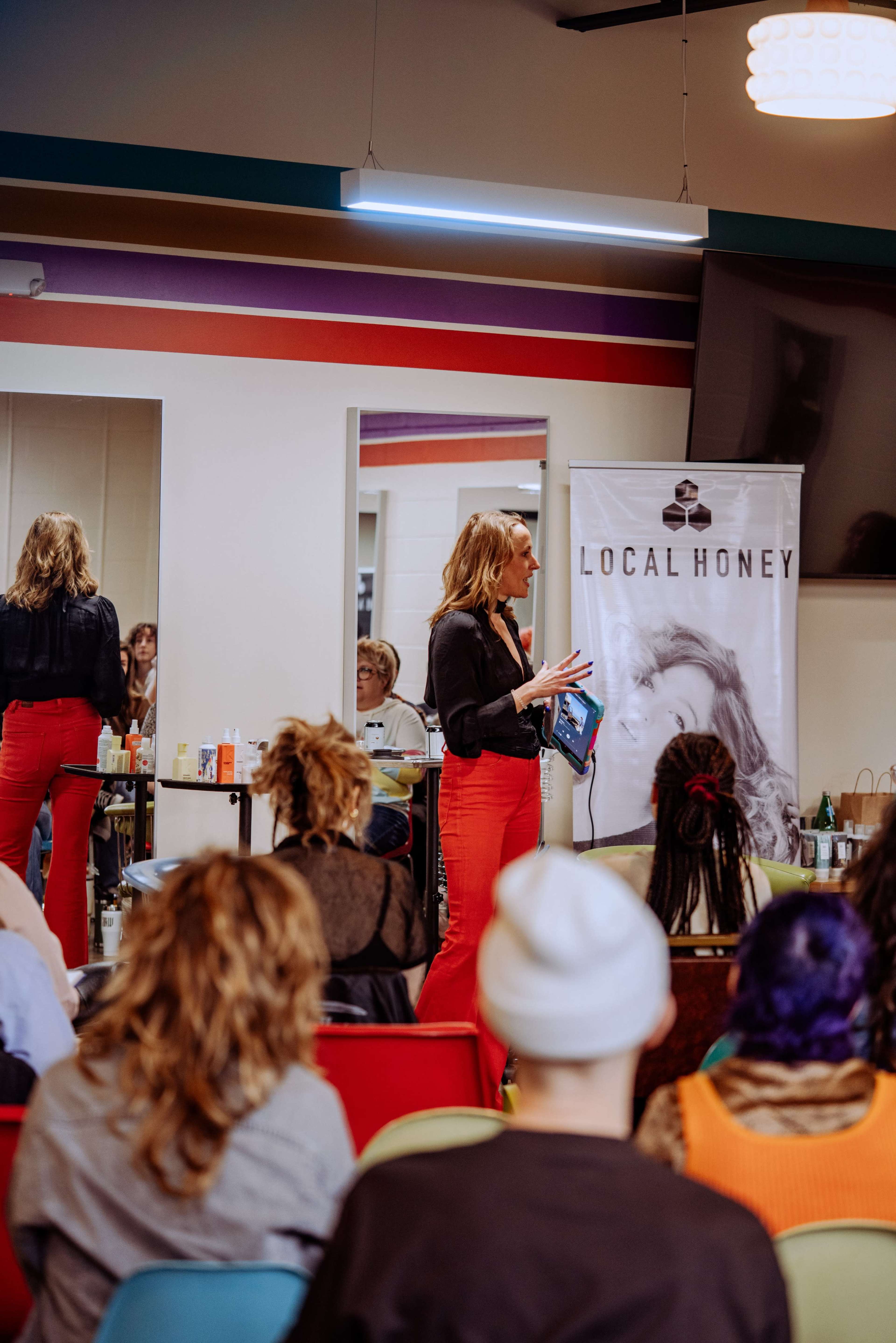 A woman in a black blouse and red pants speaks to an audience in a brightly colored room with a banner reading "LOCAL HONEY."