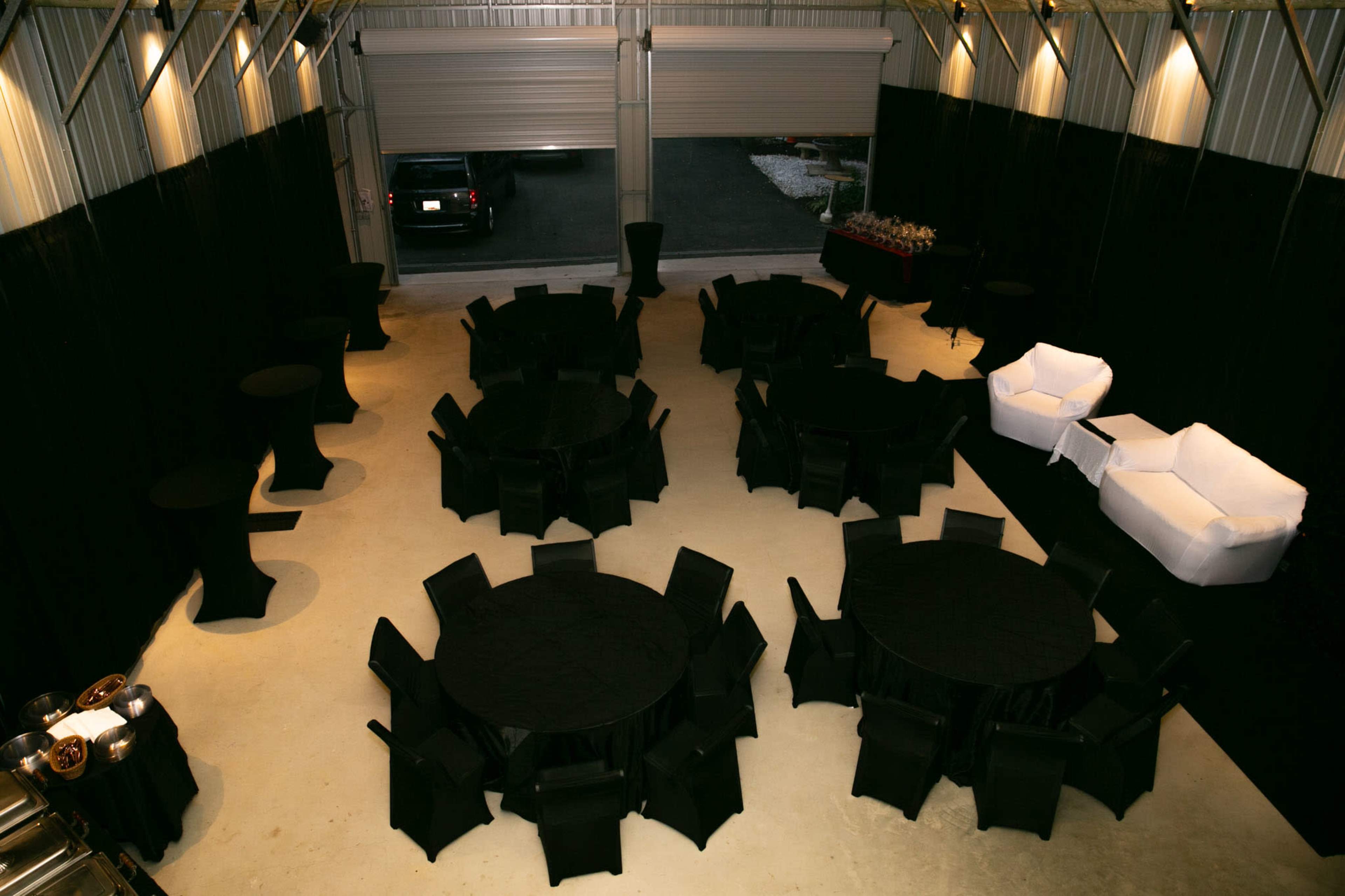 The image shows a large event space with several round black tables and chairs arranged in a circular pattern, along with two white sofas against the wall.