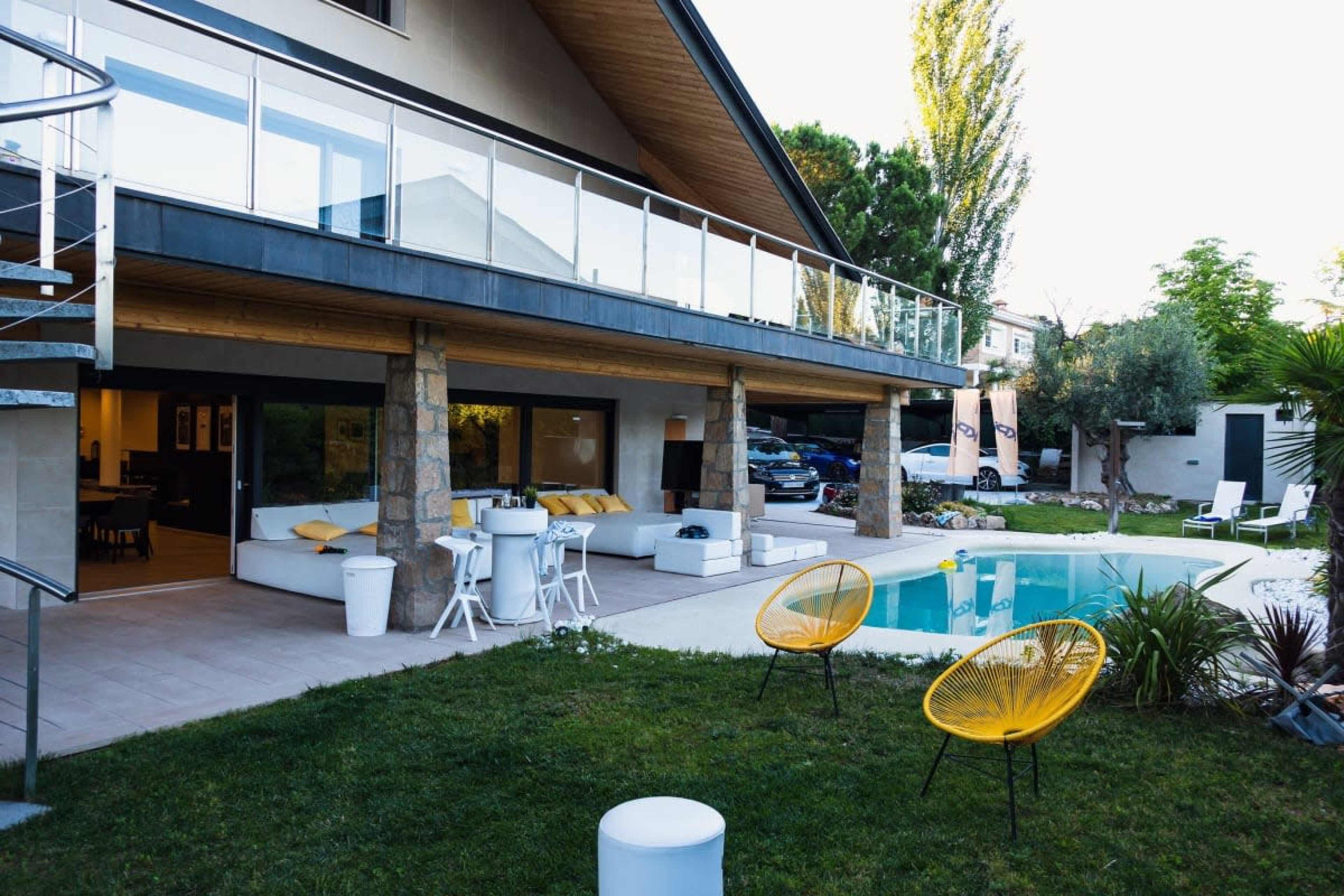 The image shows a modern house with a balcony, a pool in the backyard, and outdoor seating areas, surrounded by grass and trees.
