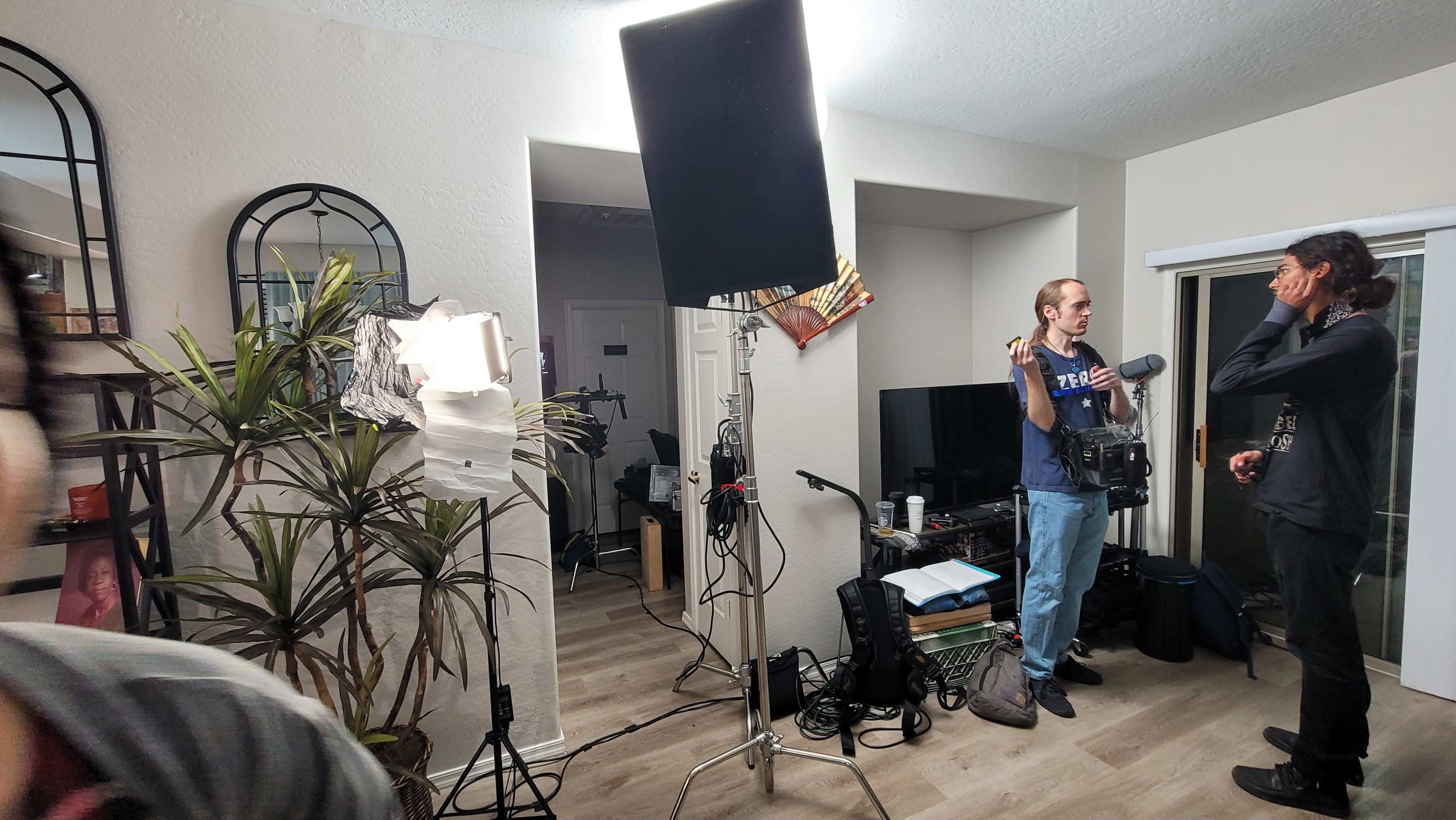 Two people are engaged in conversation in a well-lit room with various lighting equipment and plants.