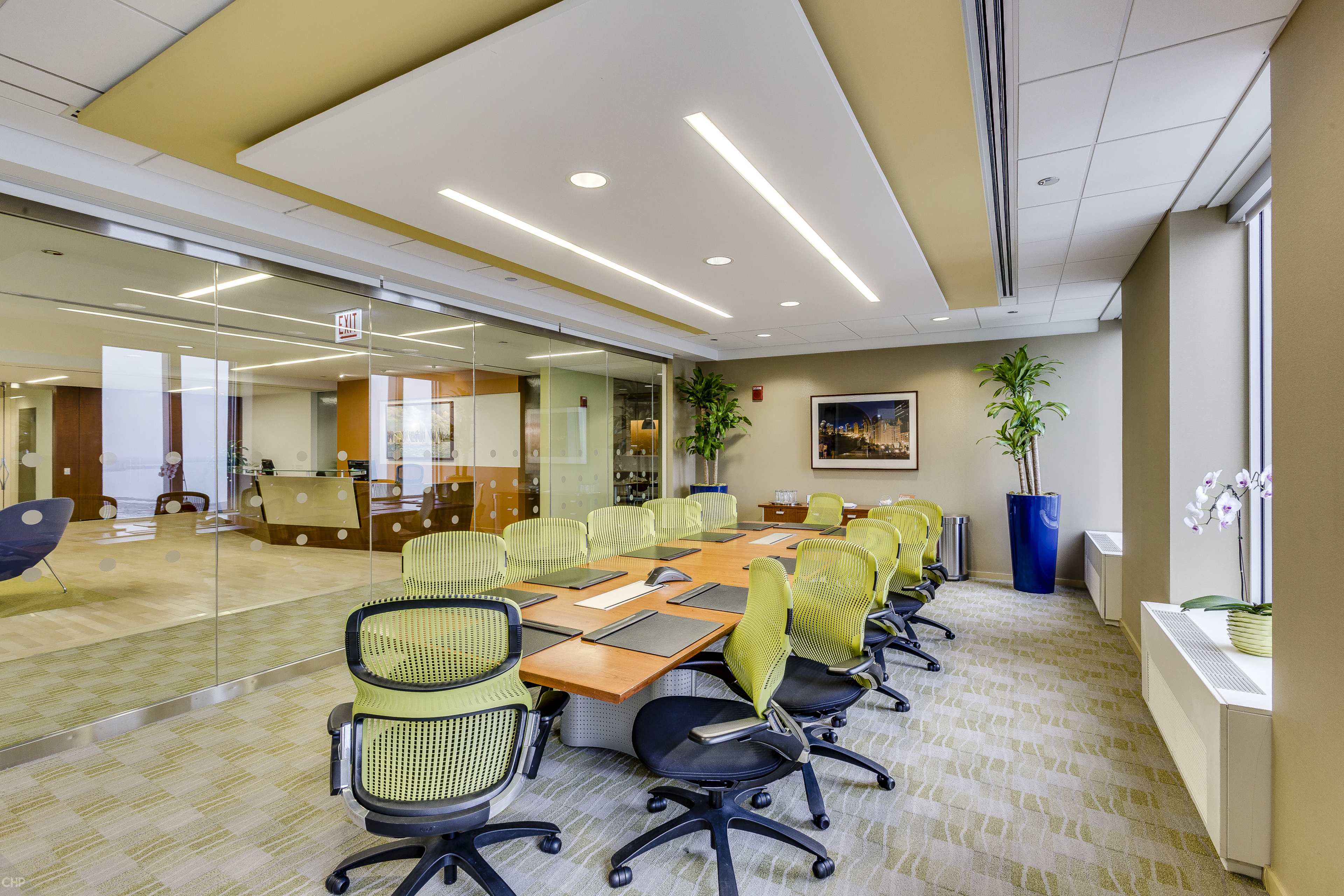A modern conference room features a long wooden table surrounded by green mesh chairs, with large windows and plants enhancing the space.
