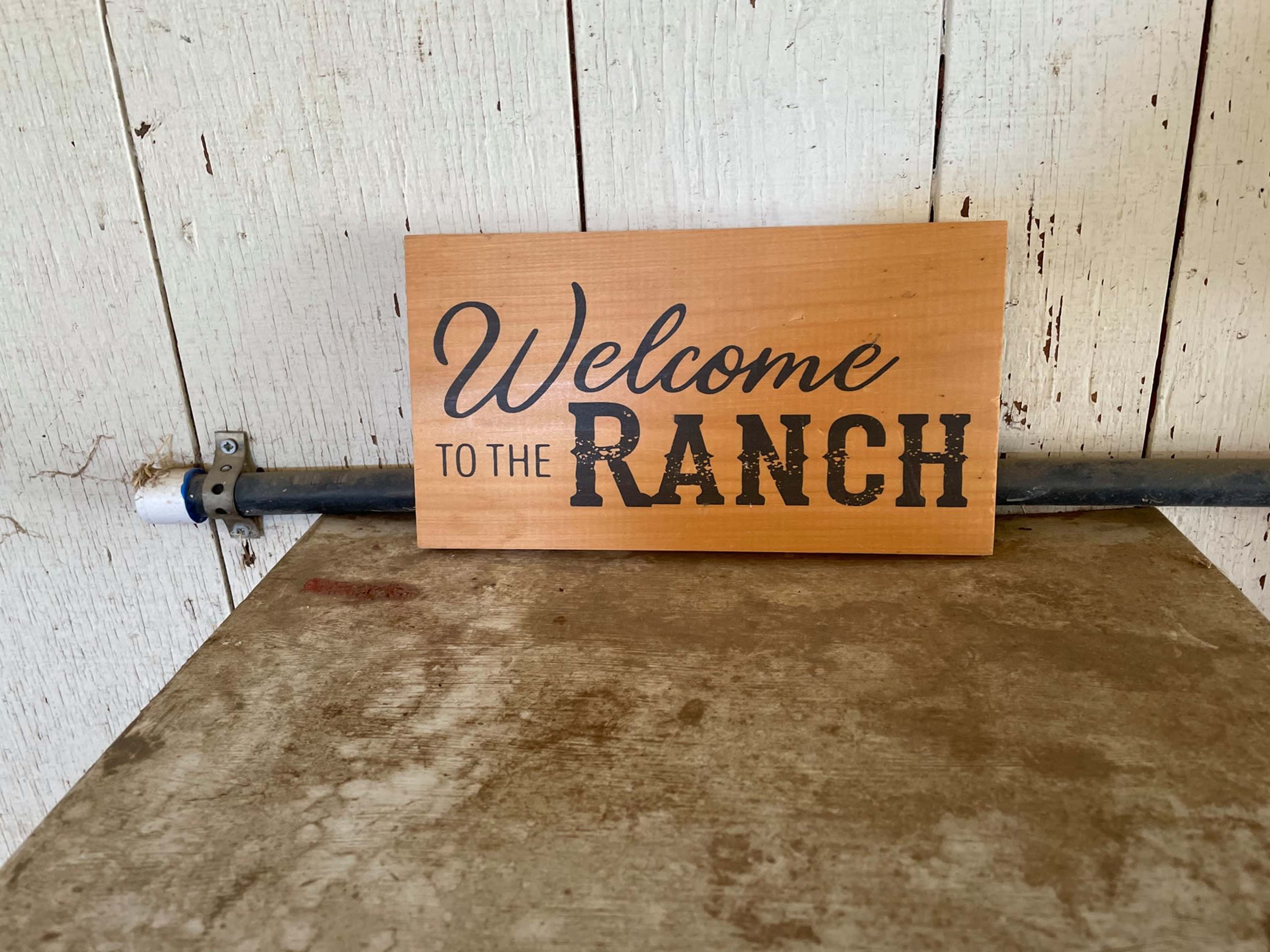 A wooden sign reading "Welcome to the Ranch" is placed on a weathered surface against a wooden wall.