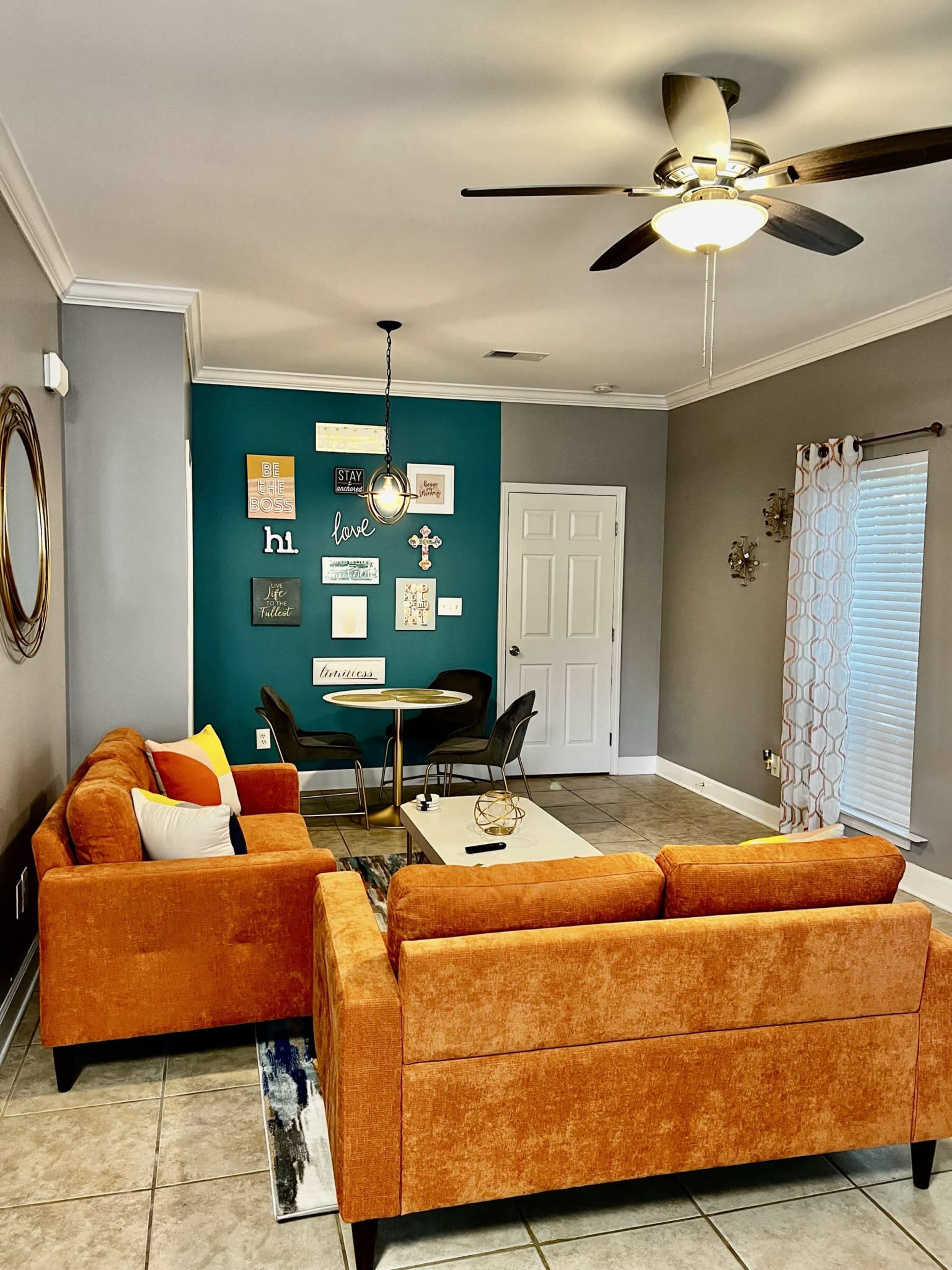 The image shows a living room with orange sofas facing a dining area, a teal accent wall adorned with framed artwork, and a ceiling fan overhead.