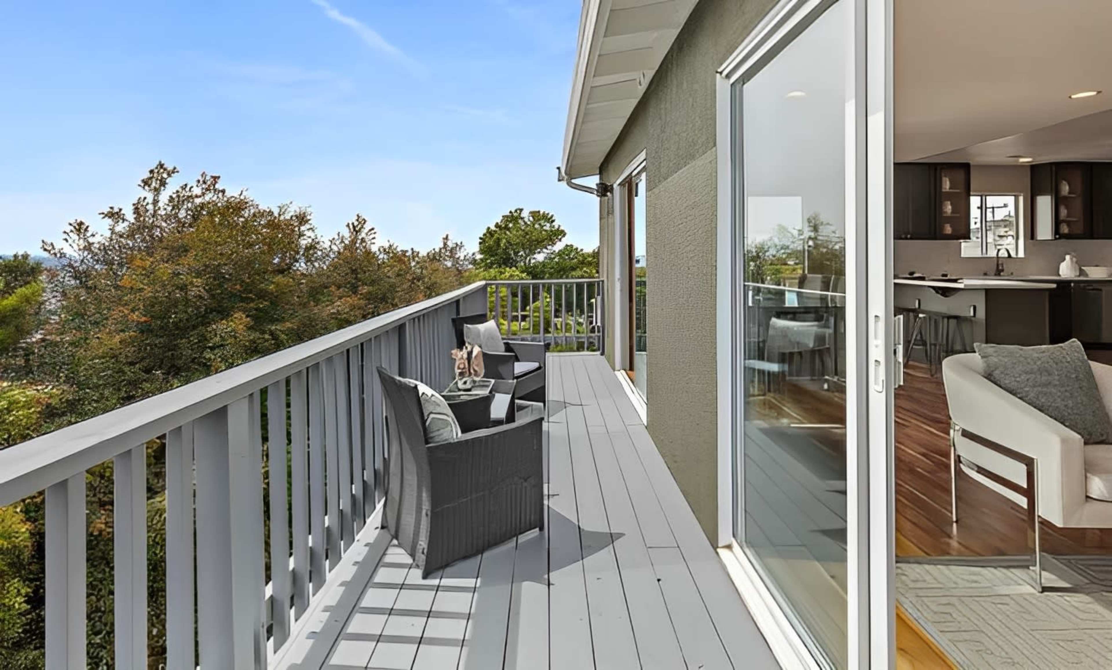 A balcony features two chairs and a small table, overlooking trees in a residential area.