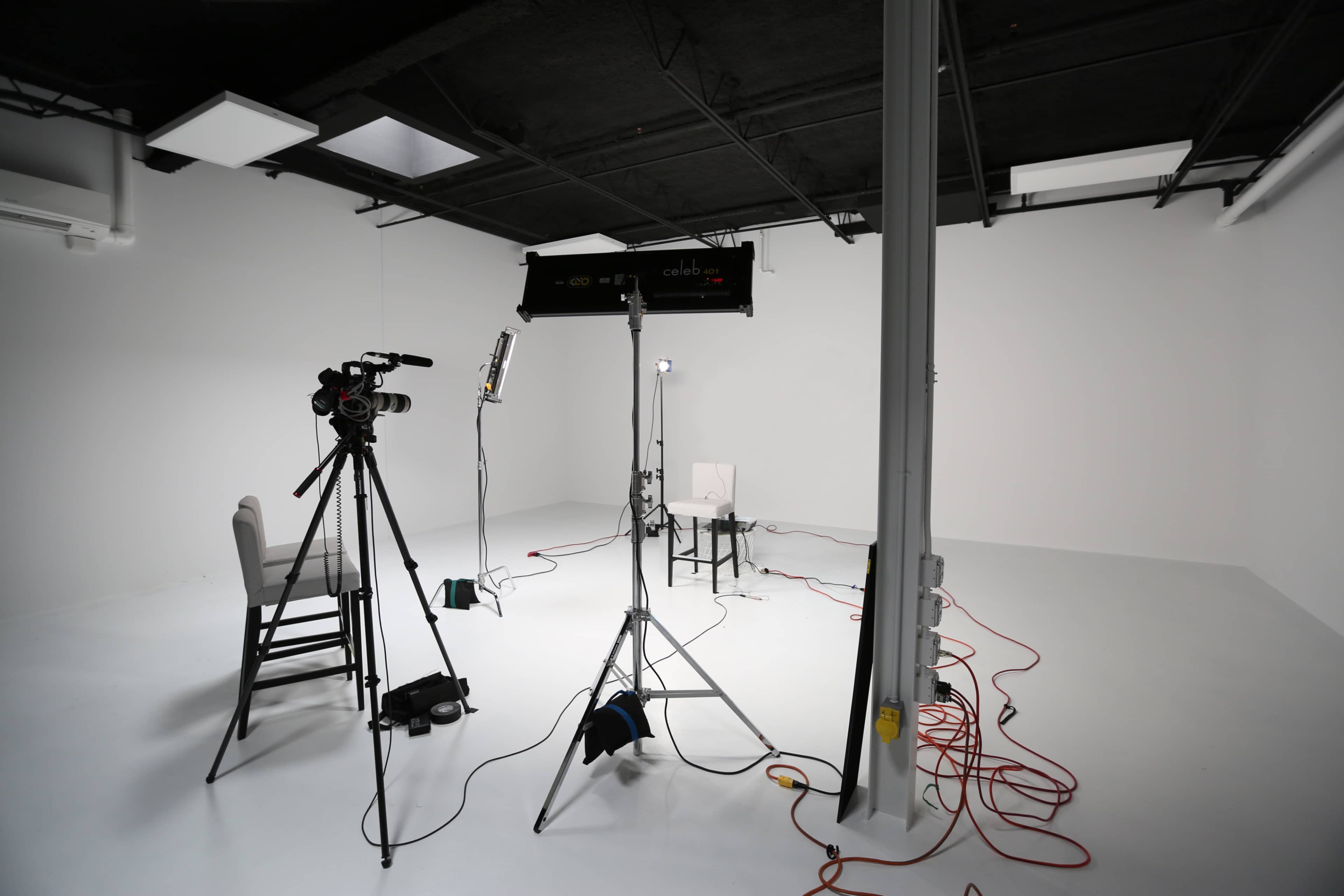 A video production setup features a camera, lighting equipment, and a single chair in an empty white studio.
