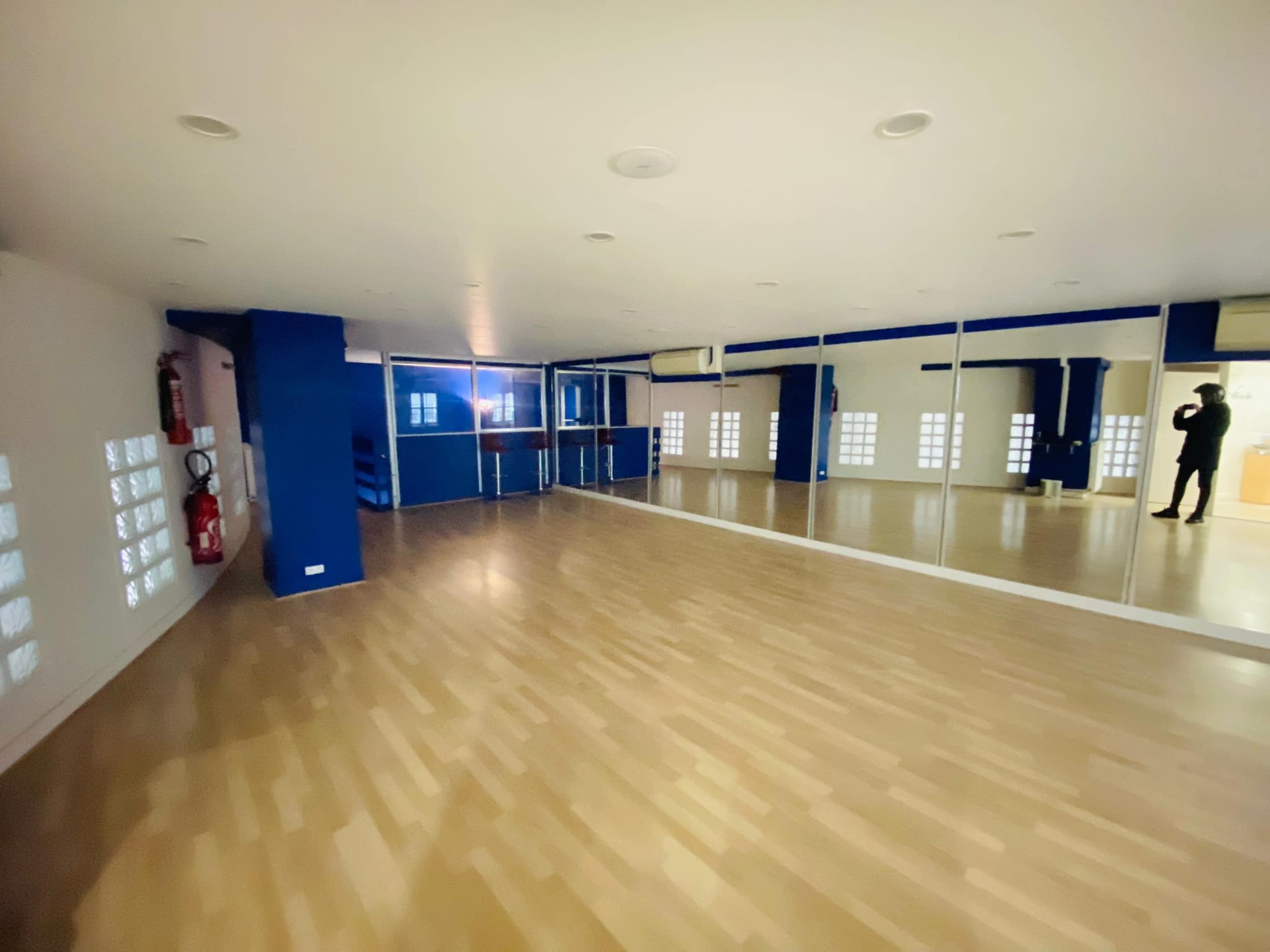 A spacious room with wooden flooring and large mirrors on one wall, featuring blue accents and a few windows.