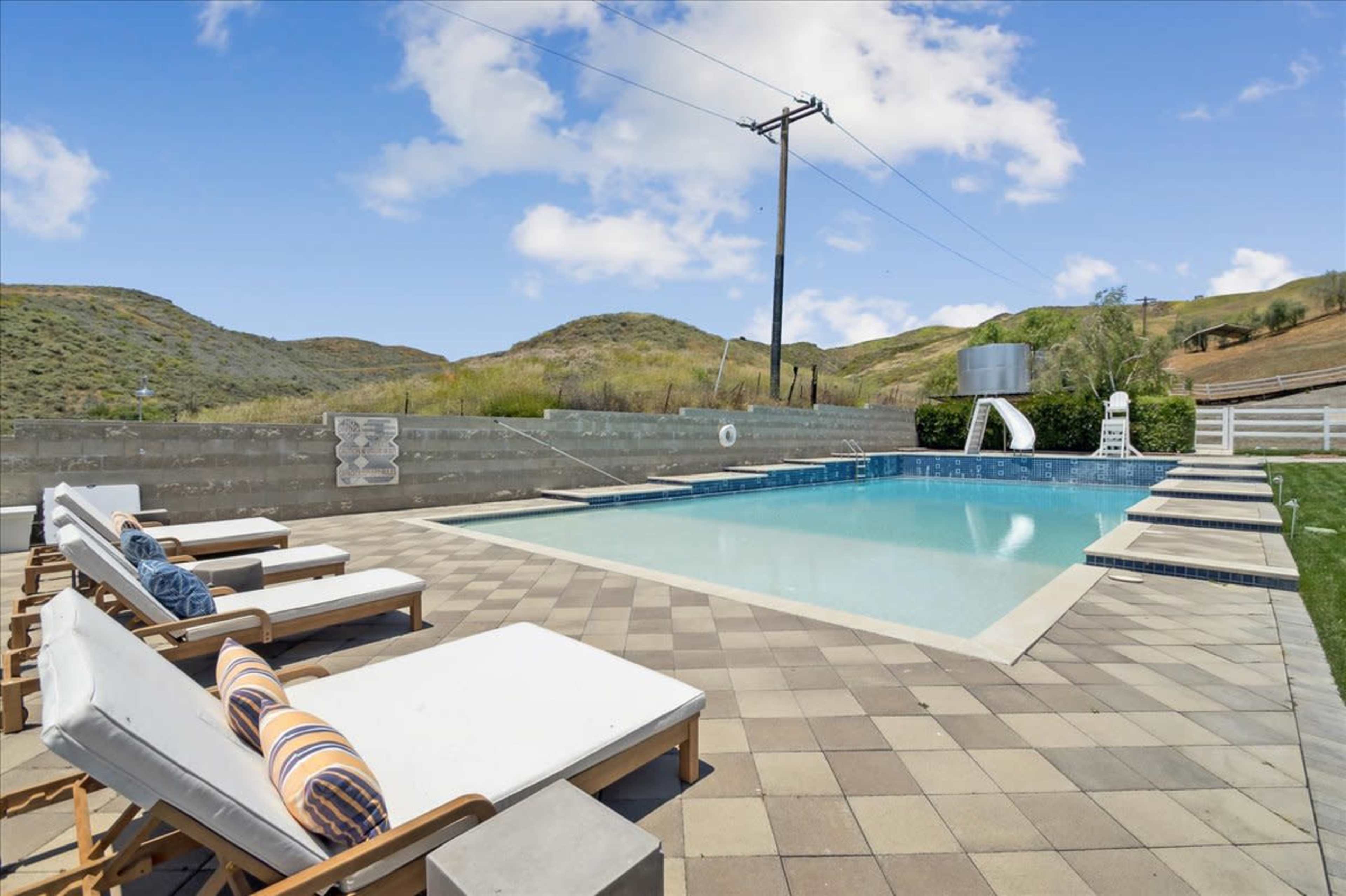 A swimming pool with tiled decking is surrounded by lounge chairs and overlooks rolling hills under a clear sky.