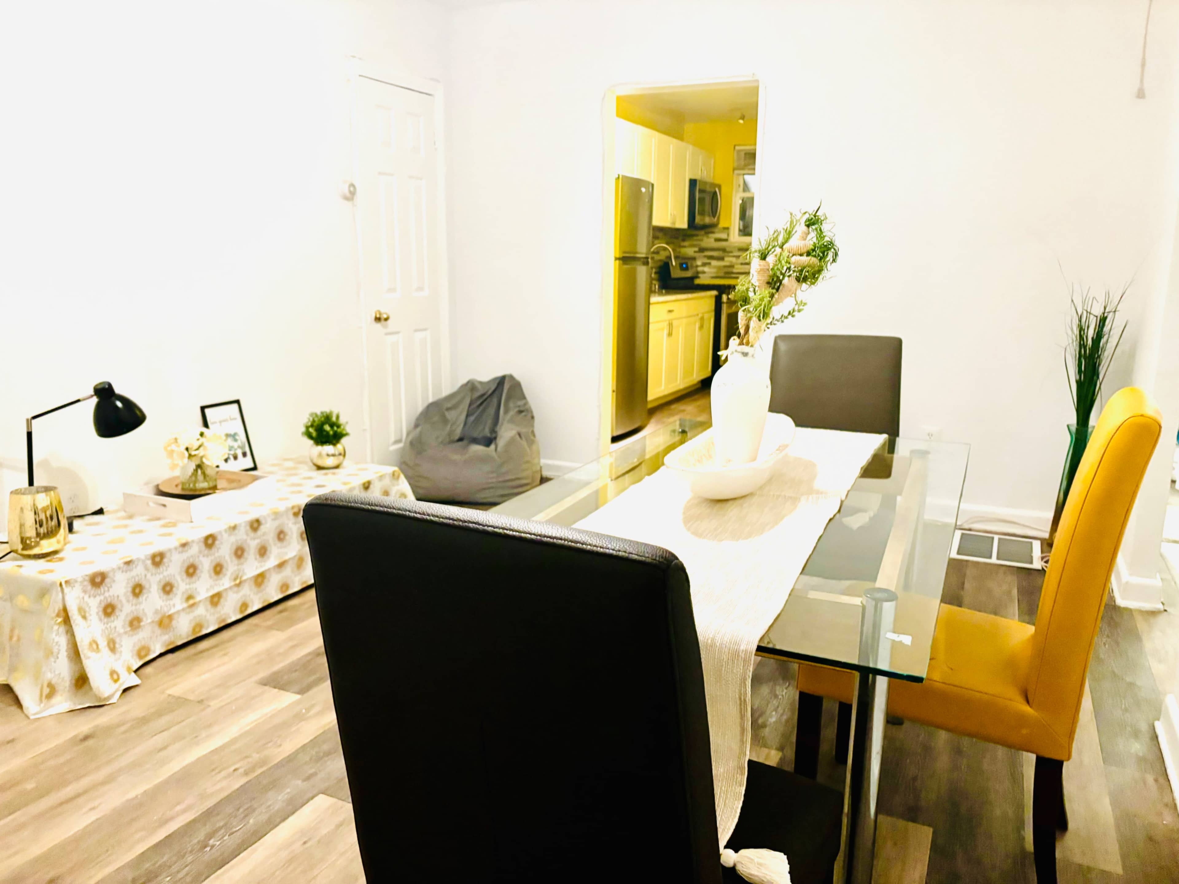 The image shows a modern dining area featuring a glass table with four chairs, a decorative table runner, and a cozy seating area with a bean bag and a plant.