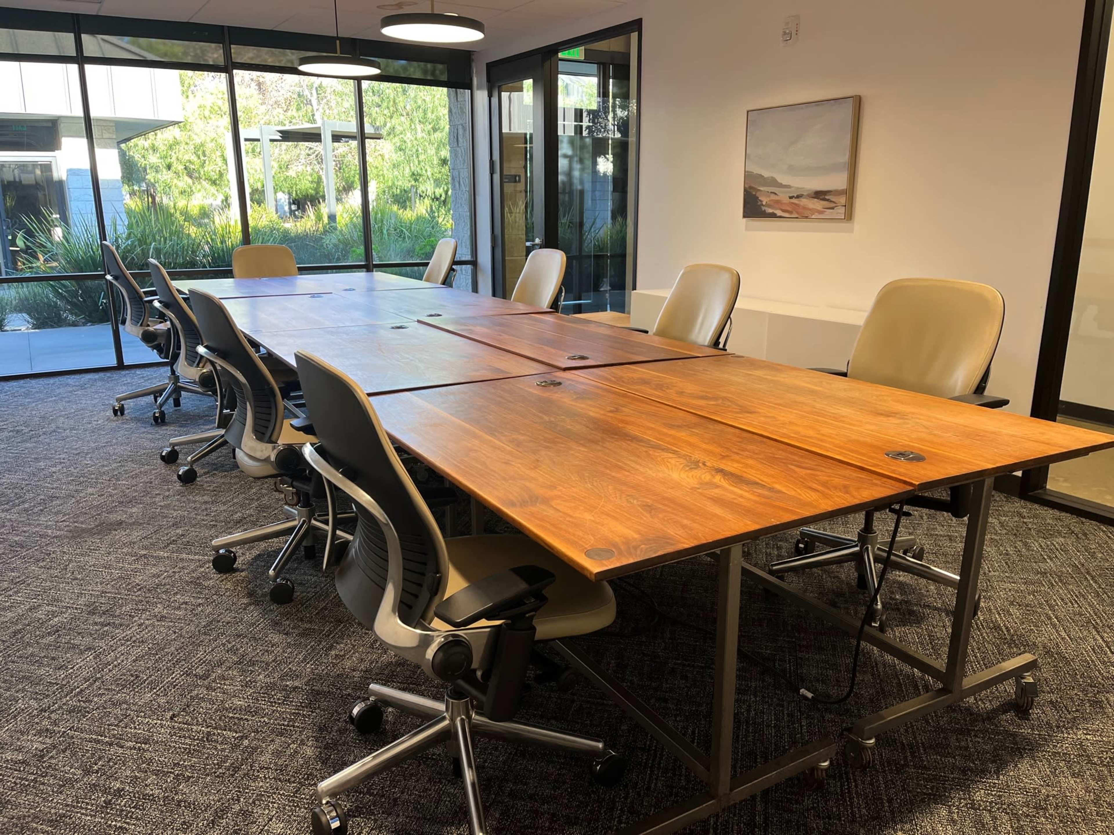 A meeting room features a long wooden table surrounded by ergonomic chairs and large windows overlooking greenery.