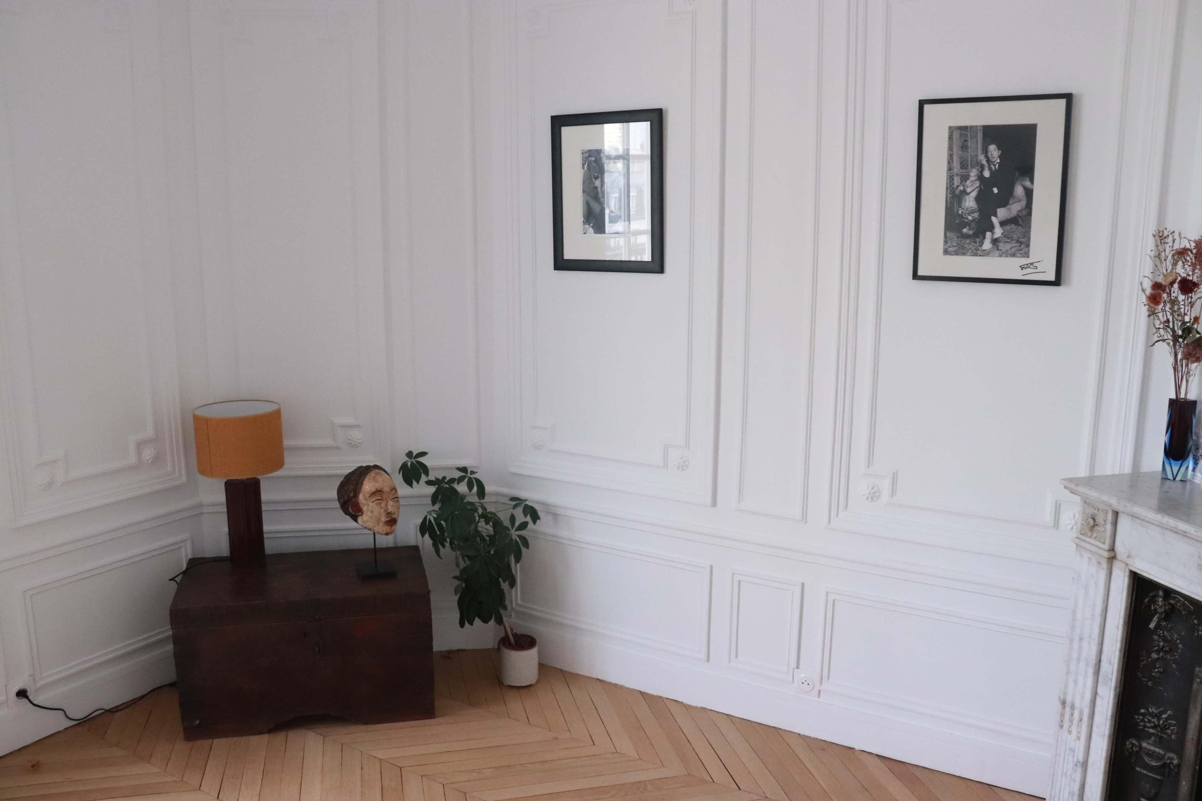 The image shows a bright room with white walls, featuring two framed pictures, a lamp on a wooden table, a small plant, and a decorative vase, all set against a herringbone wooden floor.