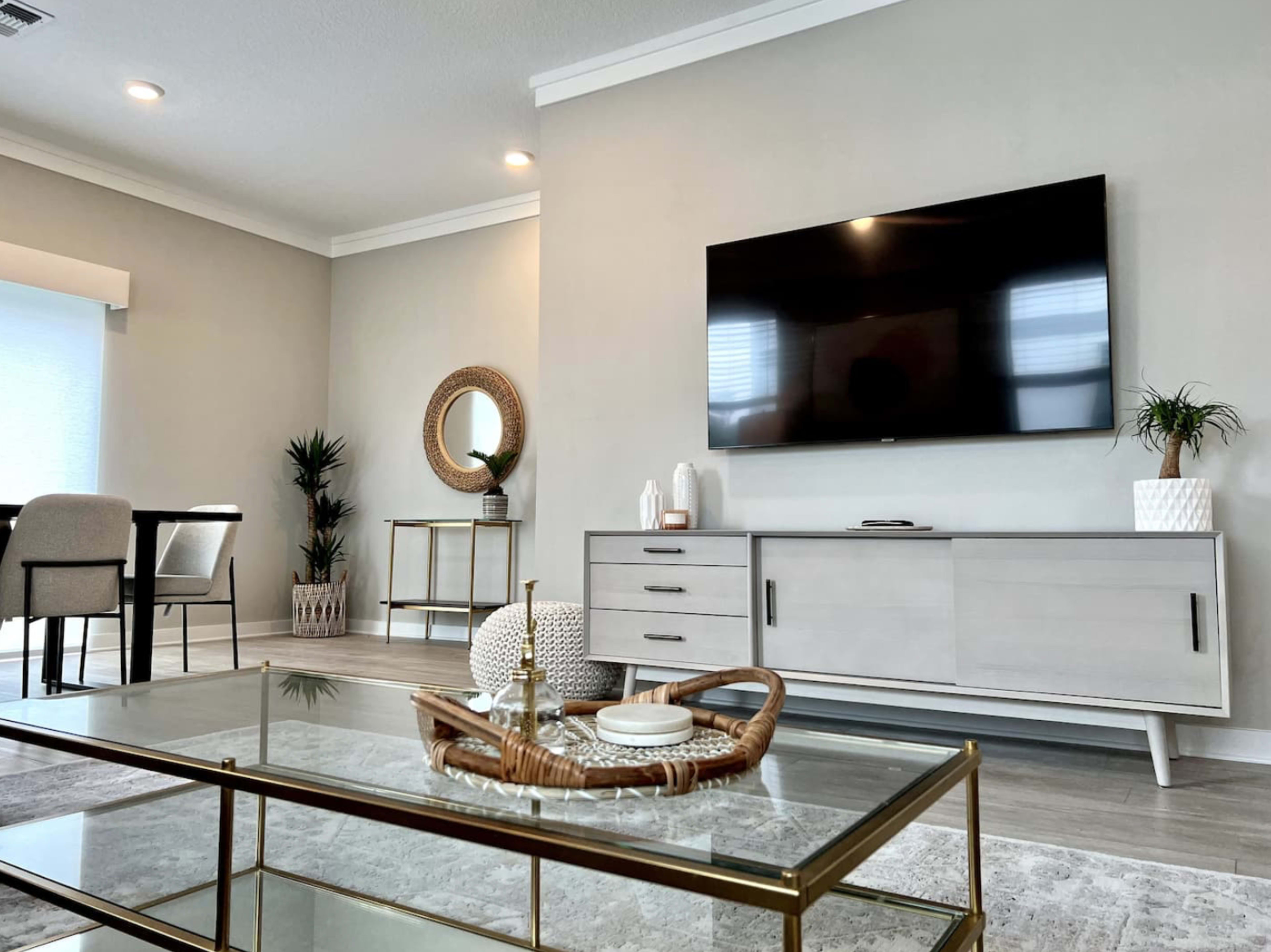 A modern living room featuring a large flat-screen TV mounted on a gray wall, a glass coffee table with decorative items, and a dining area in the background.