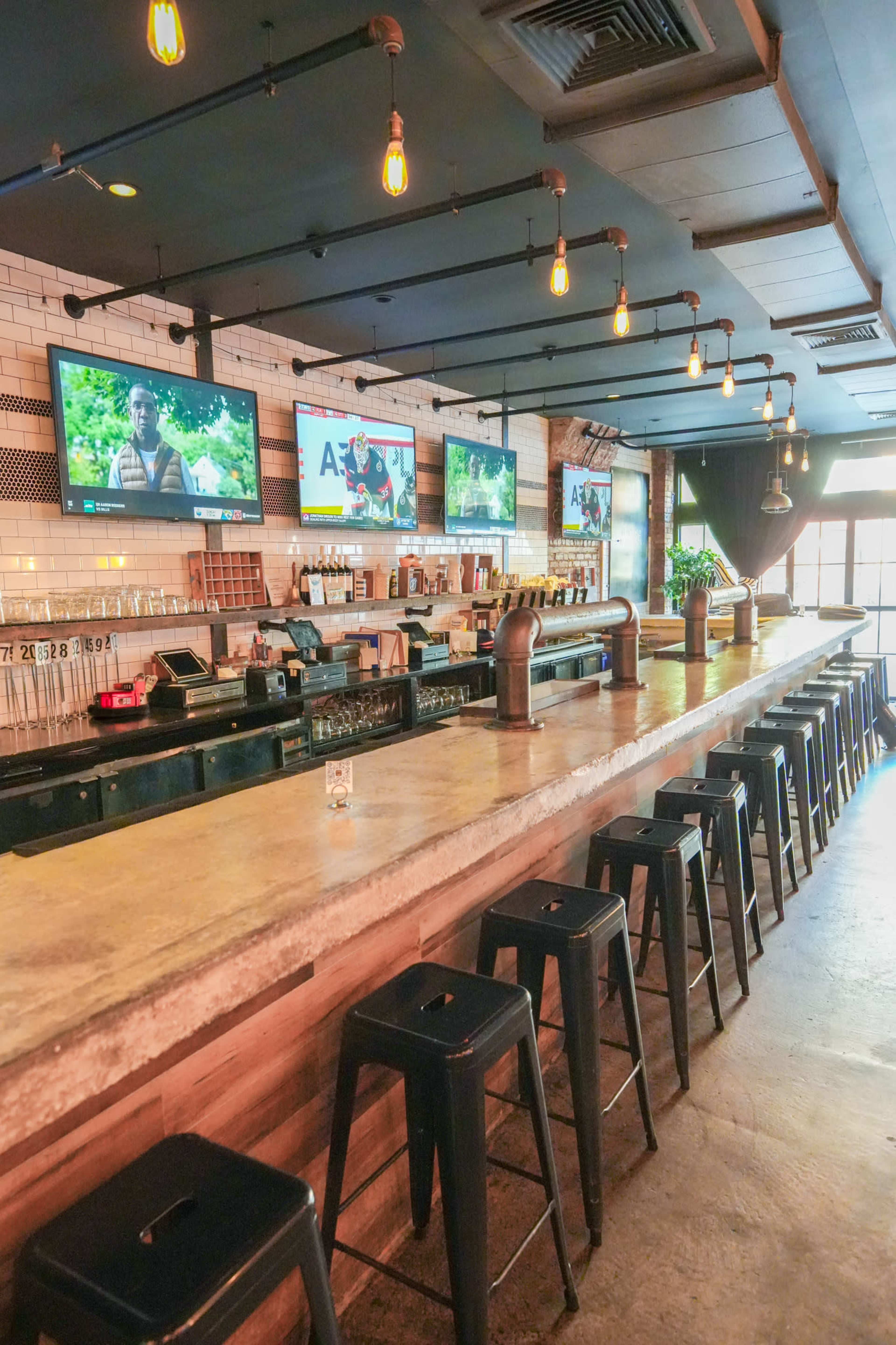 The image shows a bar interior with a long concrete countertop, metal stools, and several wall-mounted televisions displaying sports events.