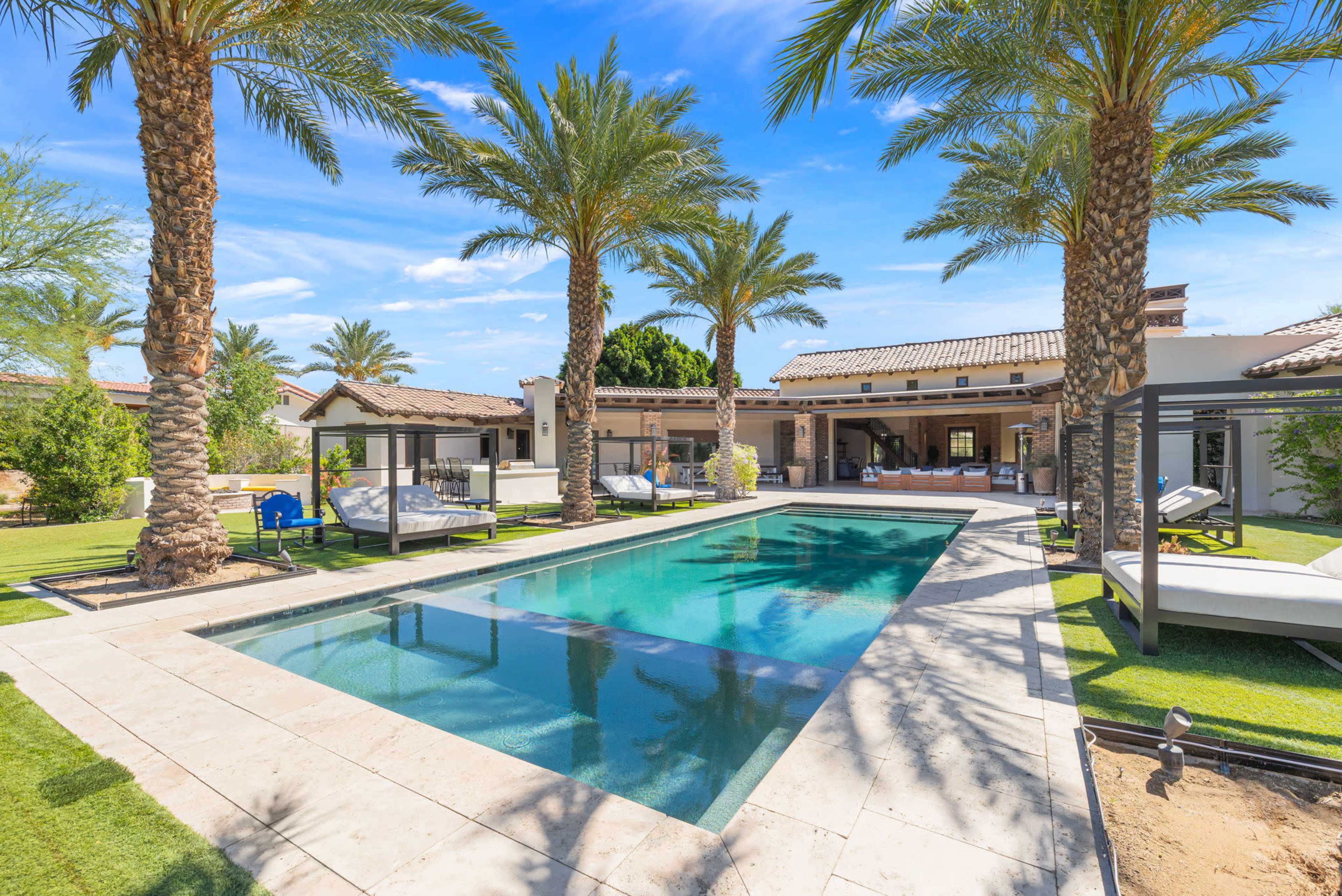 The image shows a modern backyard with a rectangular swimming pool surrounded by palm trees, lounge chairs, and a shaded seating area.