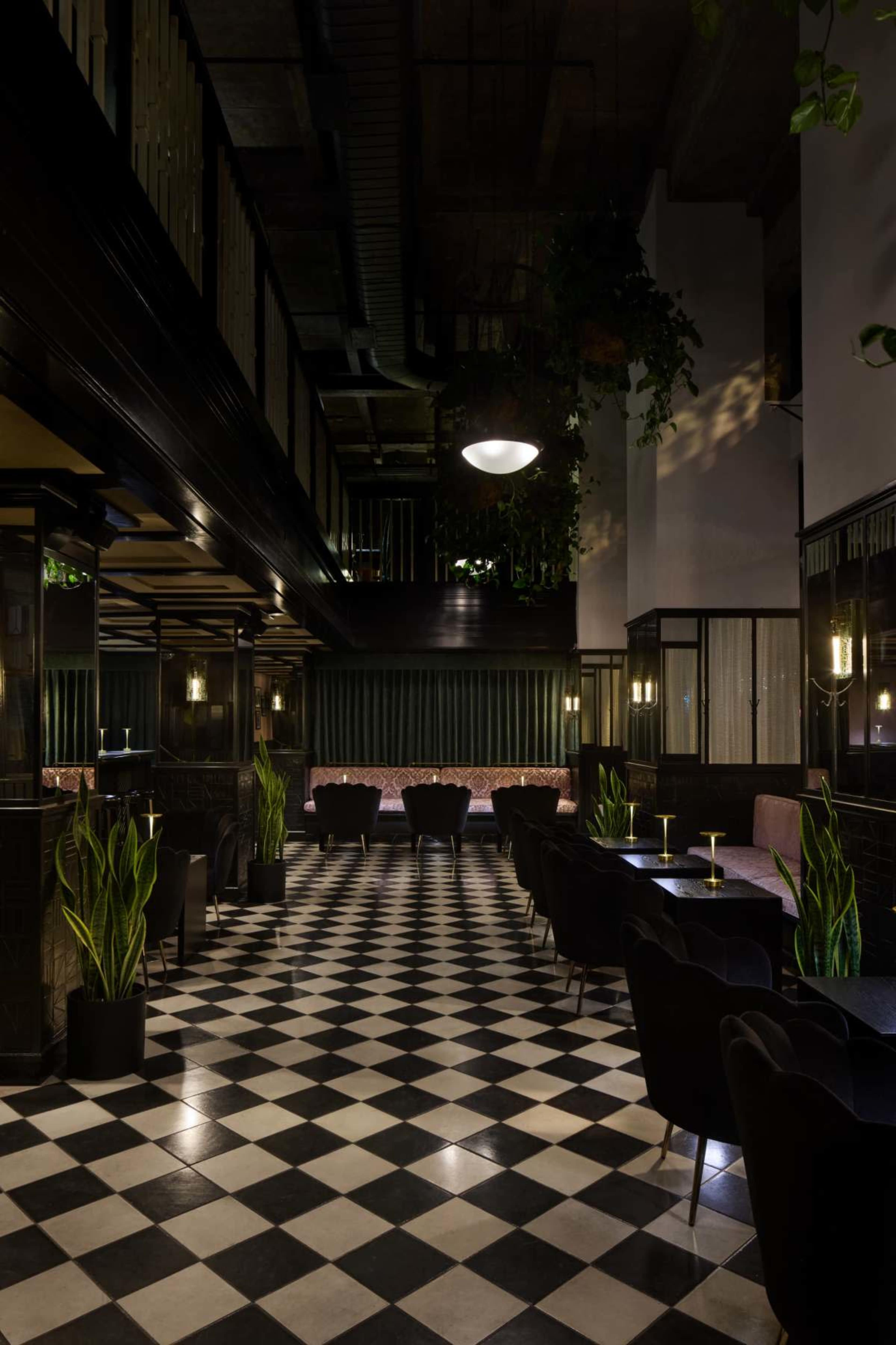 The image depicts a stylish interior of a restaurant or bar, featuring a black and white checkered floor, elegant seating arrangements, and greenery throughout the space.