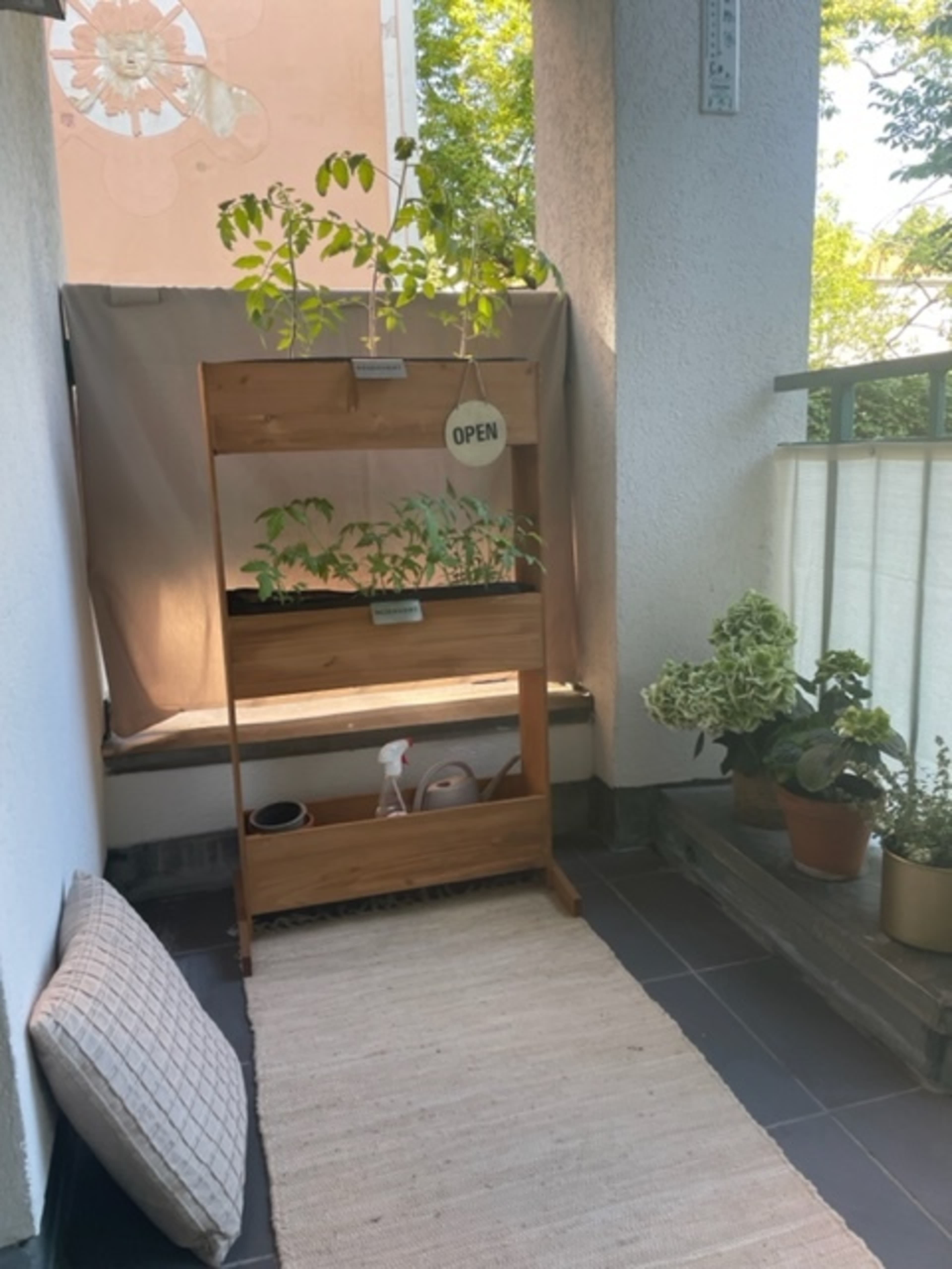 A small balcony with a wooden plant stand holding green plants, a spray bottle, and potted plants, all set against a light-colored wall.