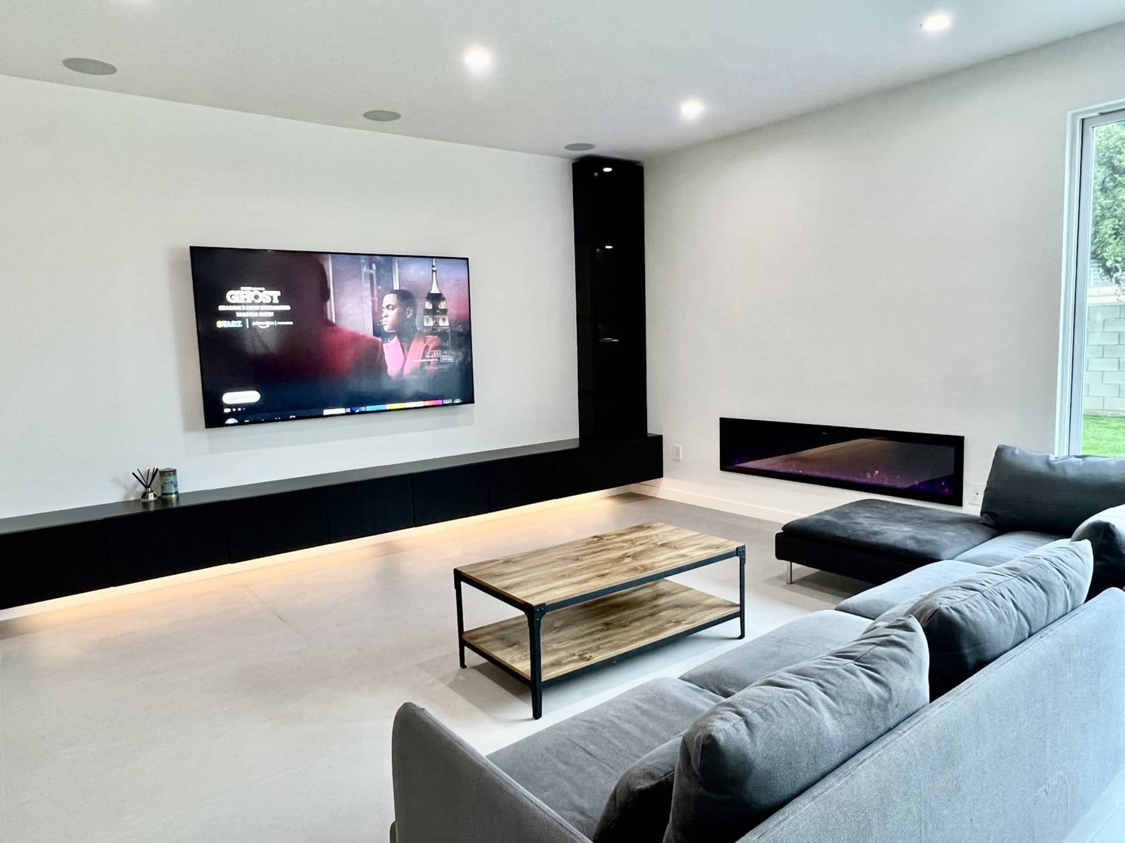 A modern living room with a large television mounted on the wall, a low coffee table, a grey sectional sofa, and a sleek electric fireplace.