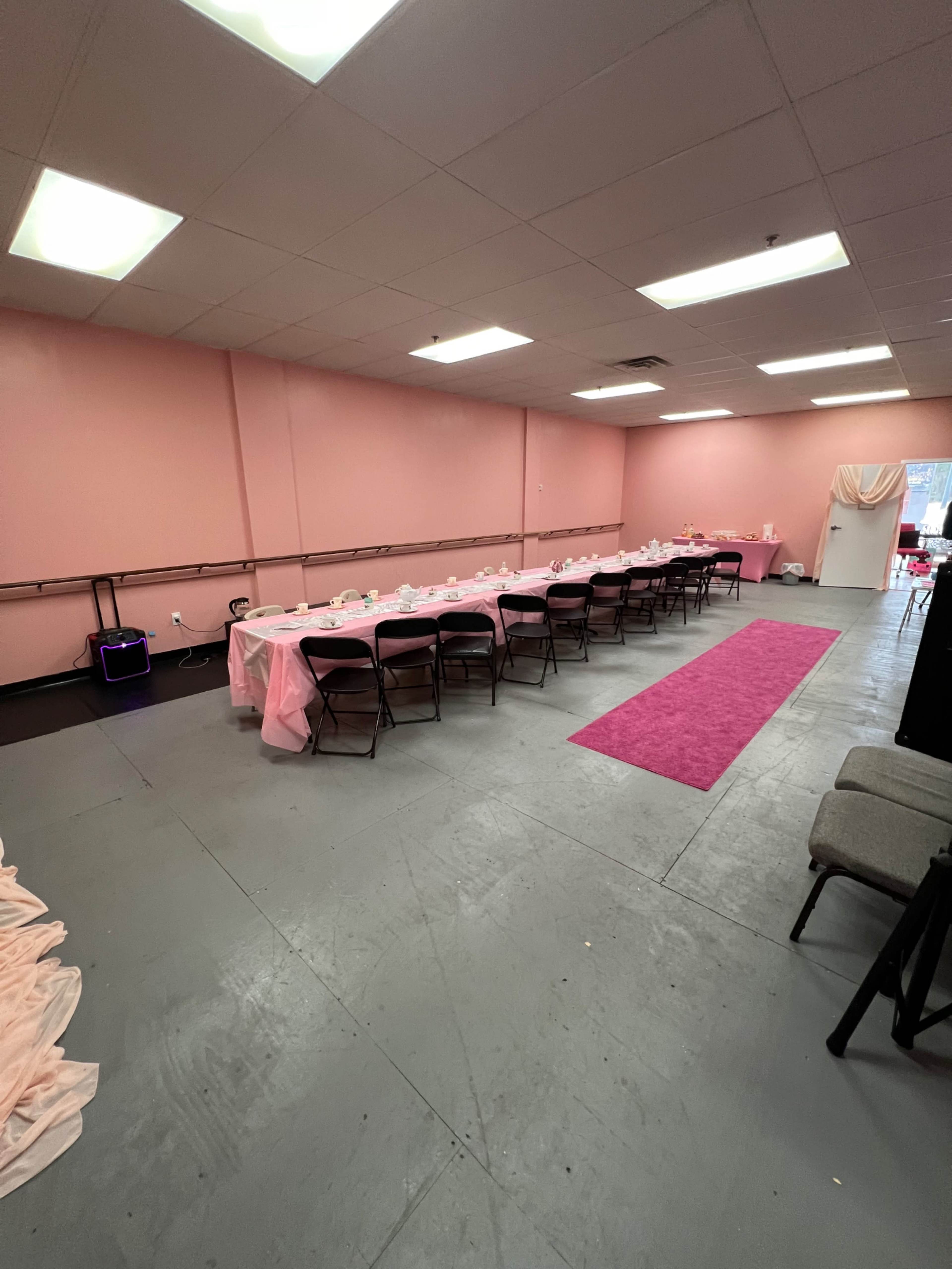 A spacious room with pink walls, featuring a long table set with a pink tablecloth and chairs, accompanied by a pink carpet running down the center.