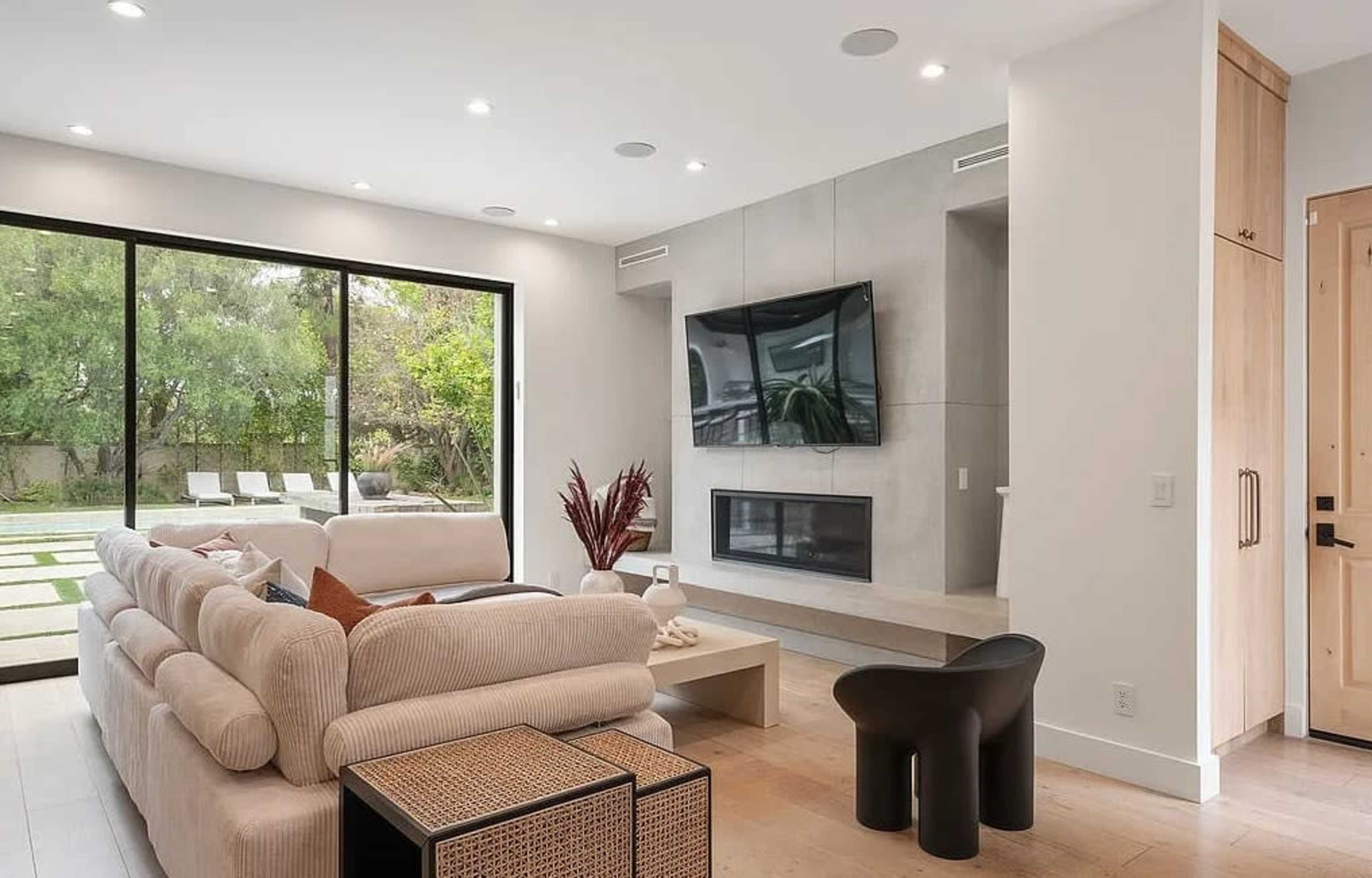 The image shows a modern living room with a large window, a minimalist design, a television mounted on the wall, and a cozy seating area.