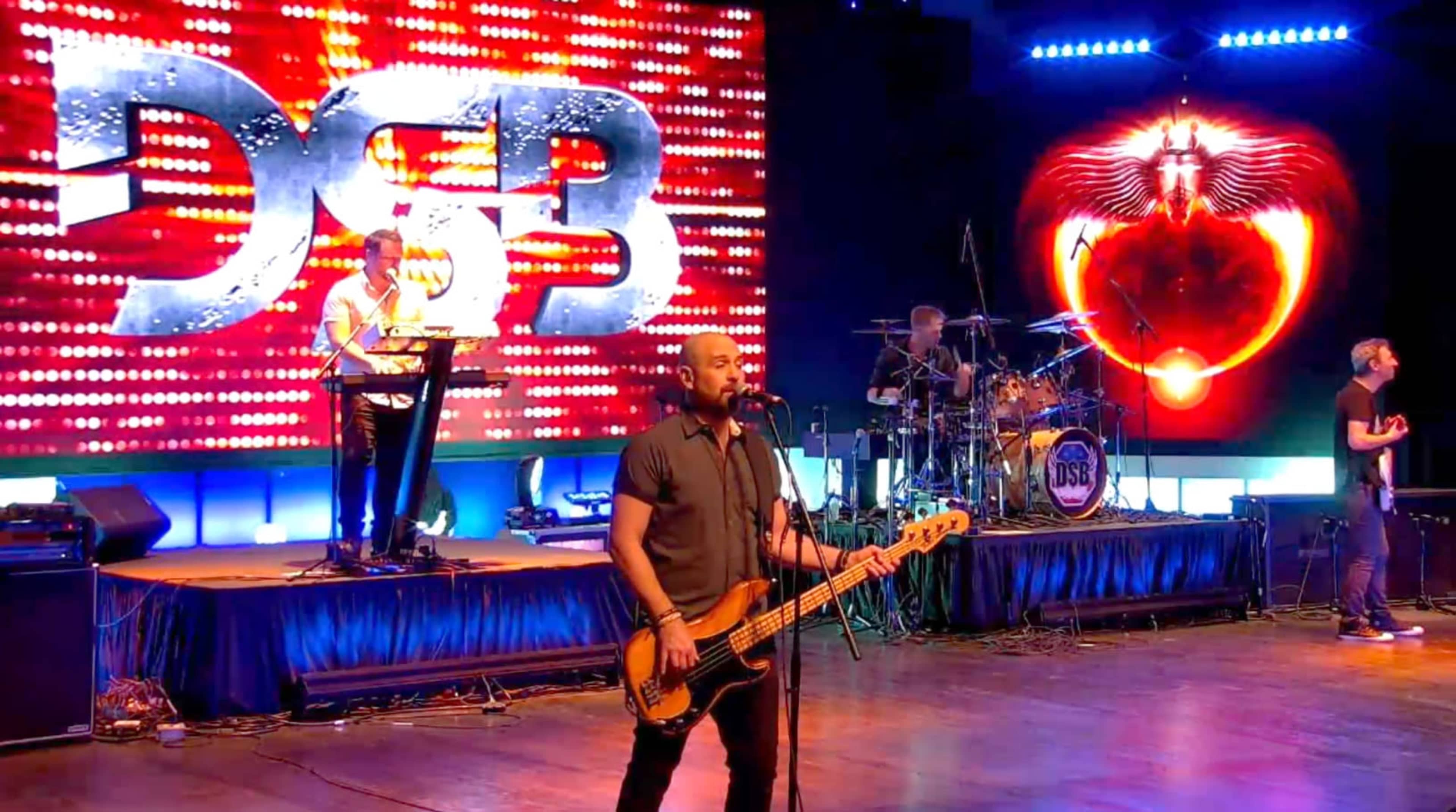 A band performs on stage with a vibrant backdrop featuring illuminated graphics and instruments.