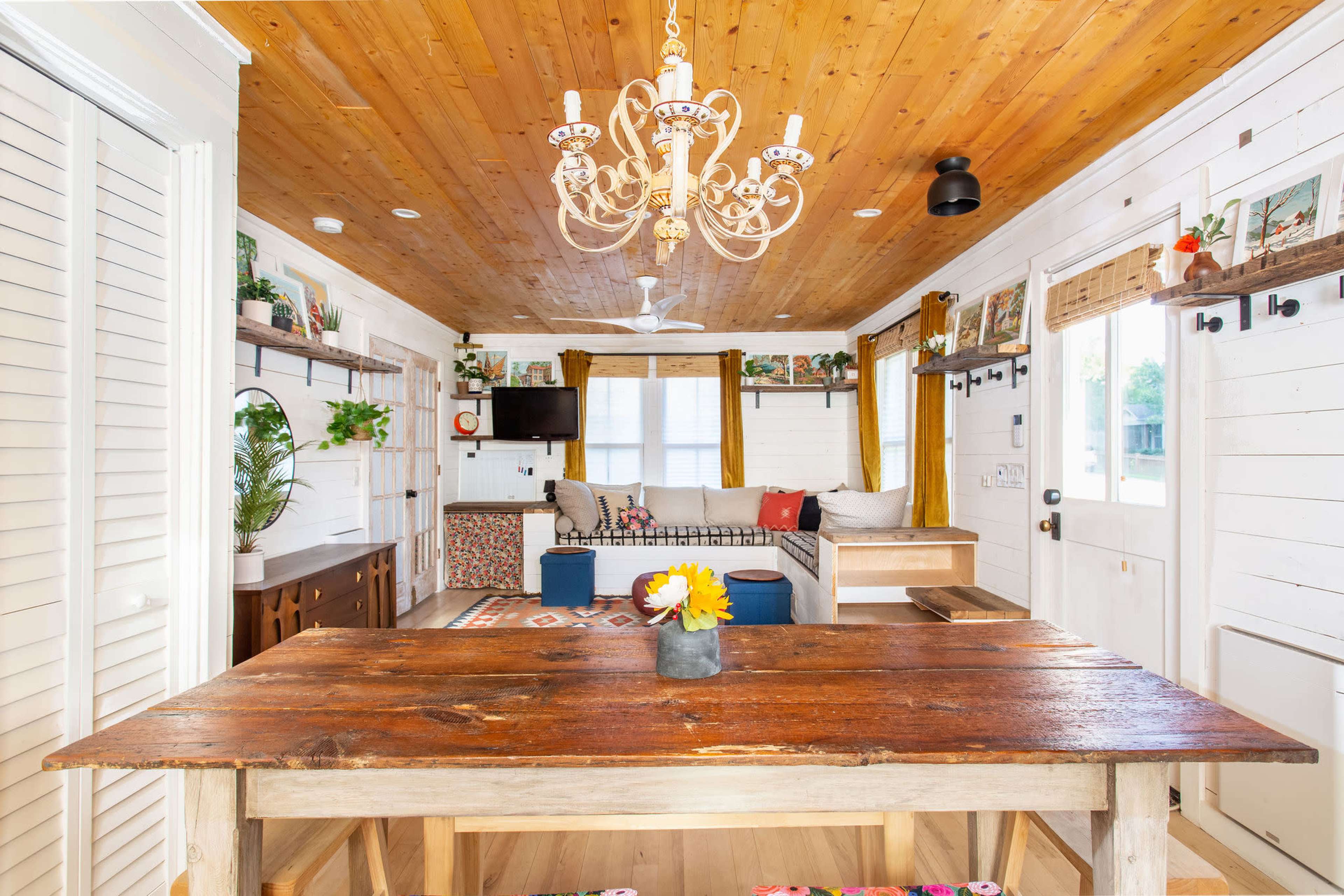 A wooden dining table with a flower vase sits in front of a brightly lit room featuring a couch, television, and potted plants.