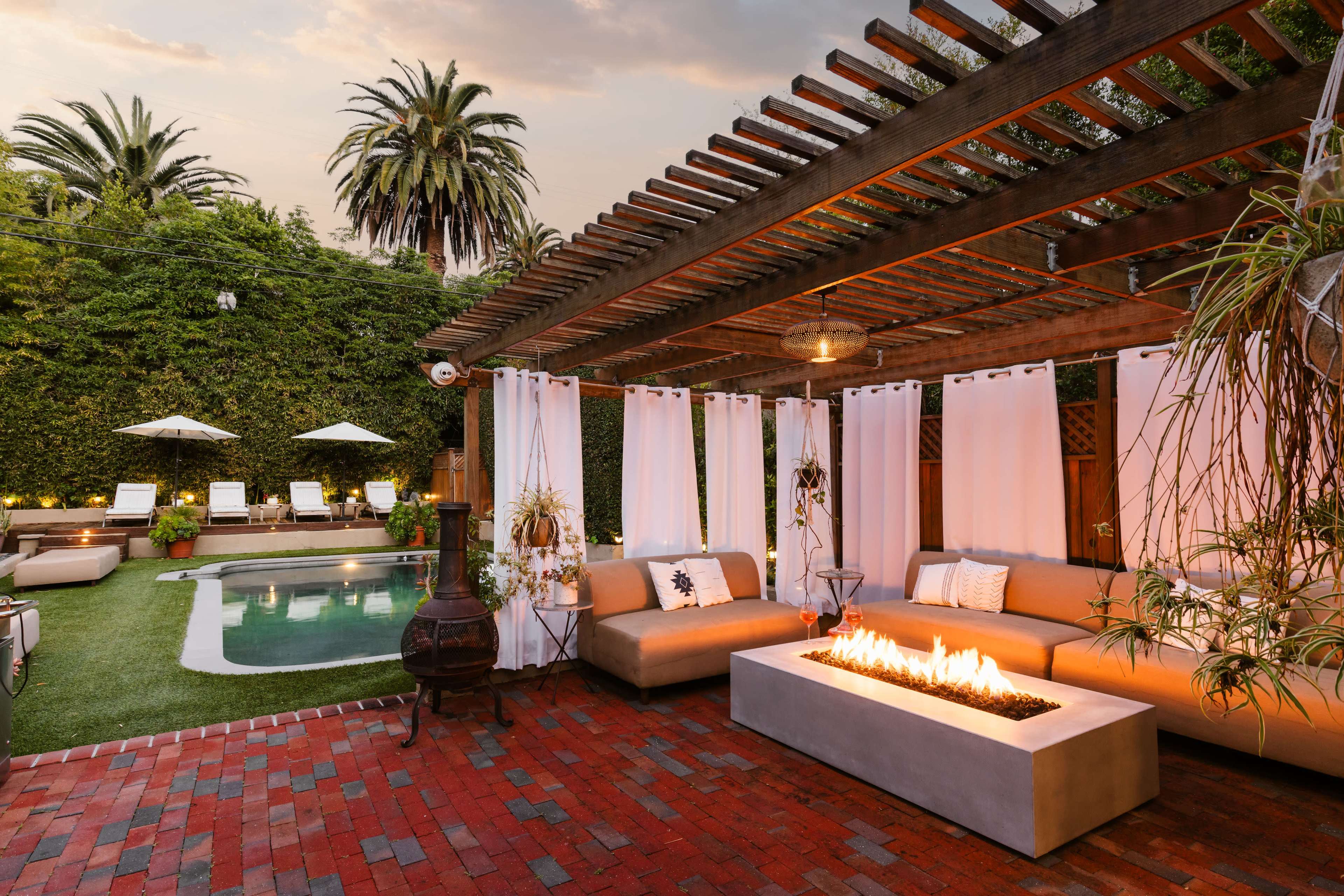A modern outdoor seating area features a fire pit, surrounded by lush greenery and a pool with lounge chairs under umbrellas in the background.