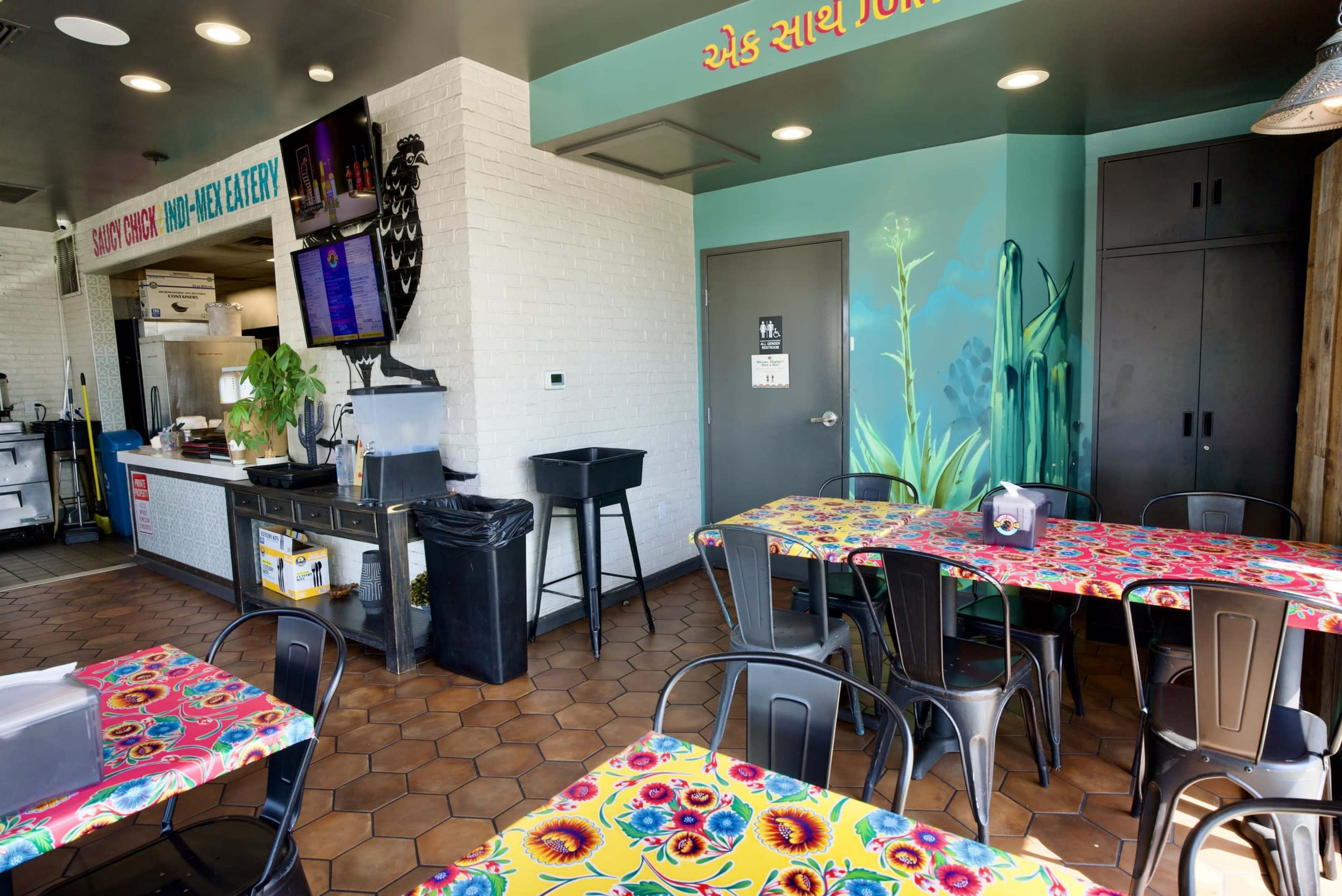 The image shows a brightly colored restaurant interior with patterned tables, a serving area with a menu display, and a mural of plants on the wall.