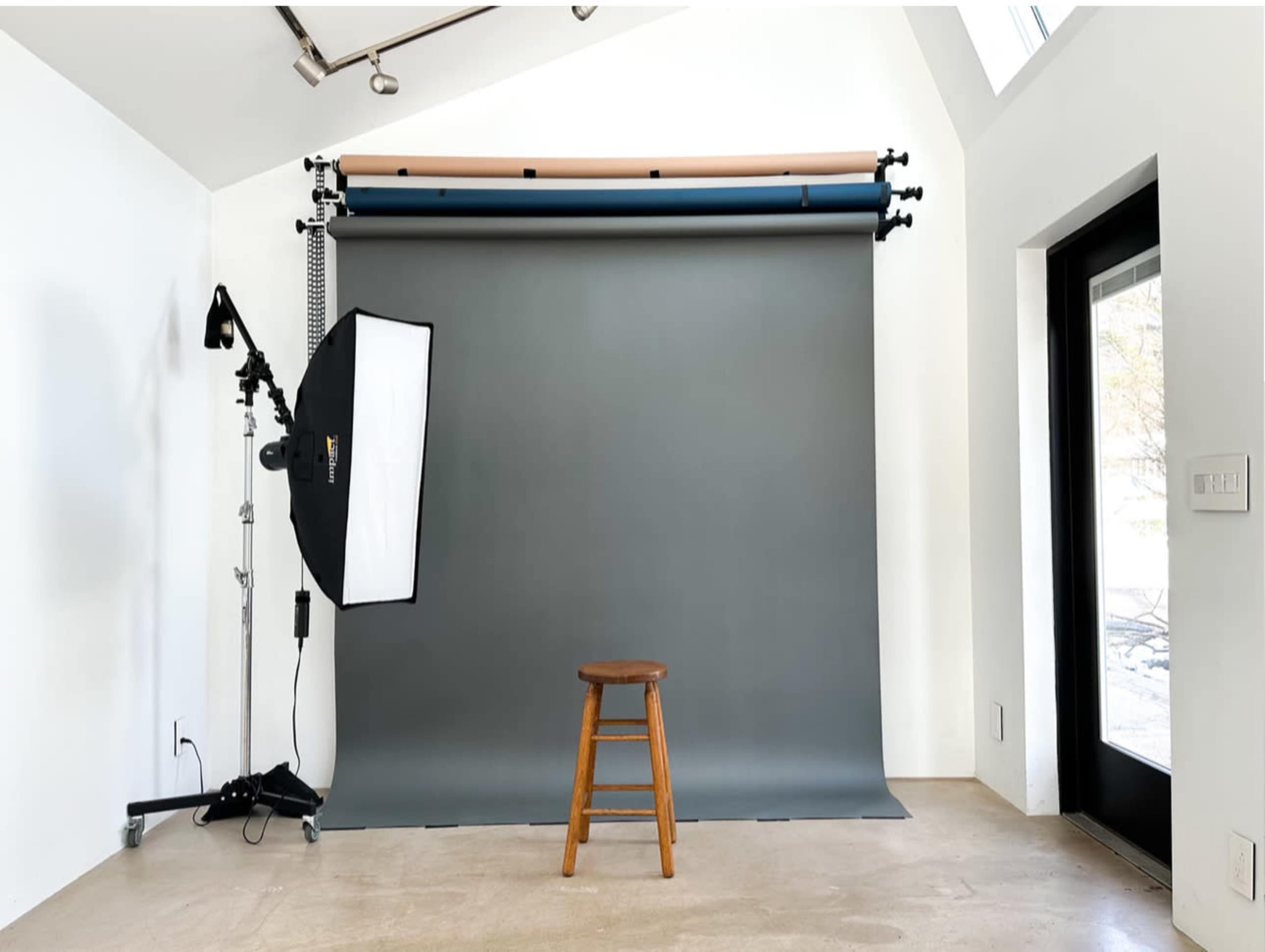 A photography studio features a gray backdrop, a wooden stool, and a lighting setup on the left side.