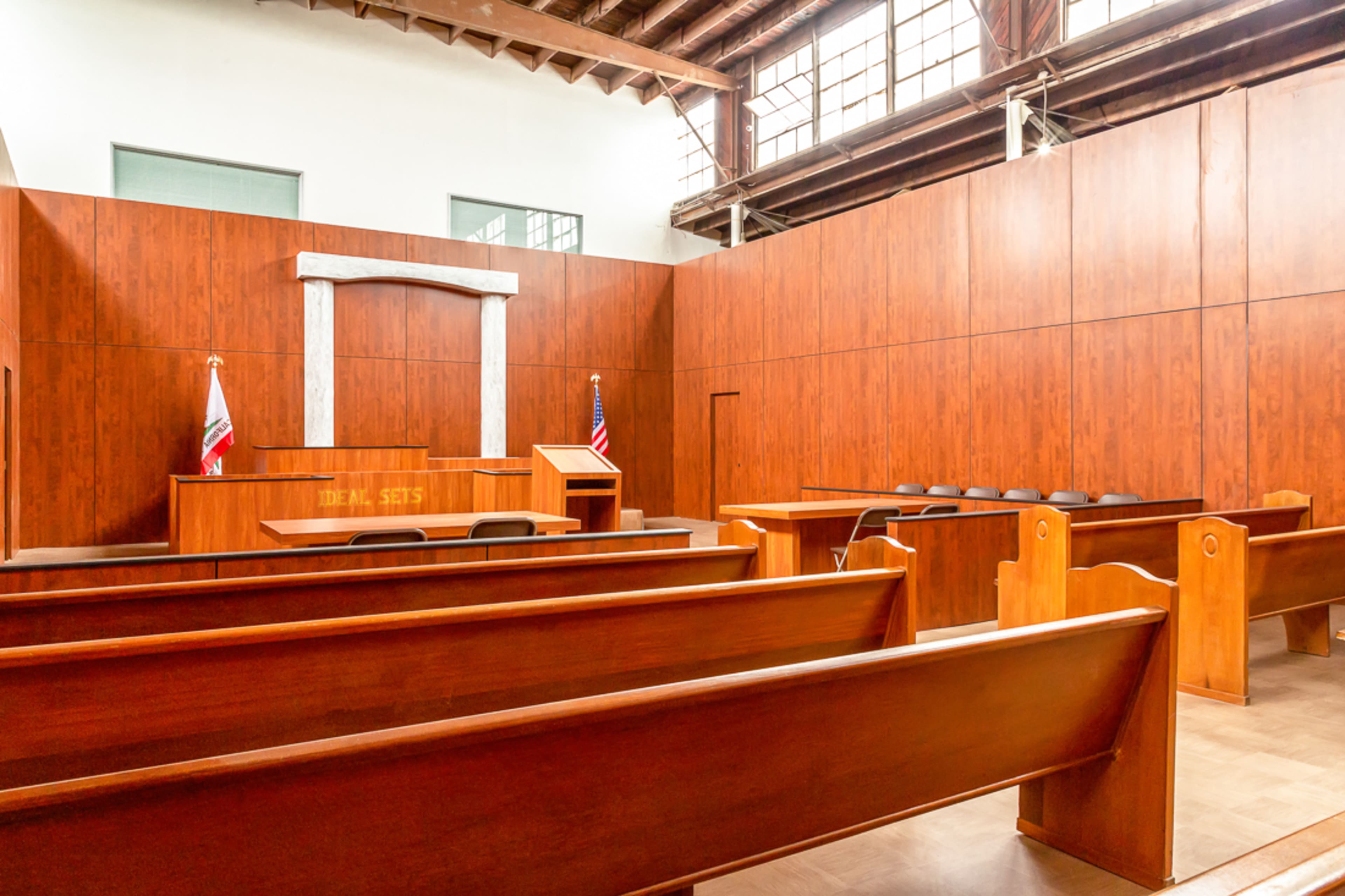 A wooden-paneled courtroom with empty benches, a judge's bench, and flags at the front.