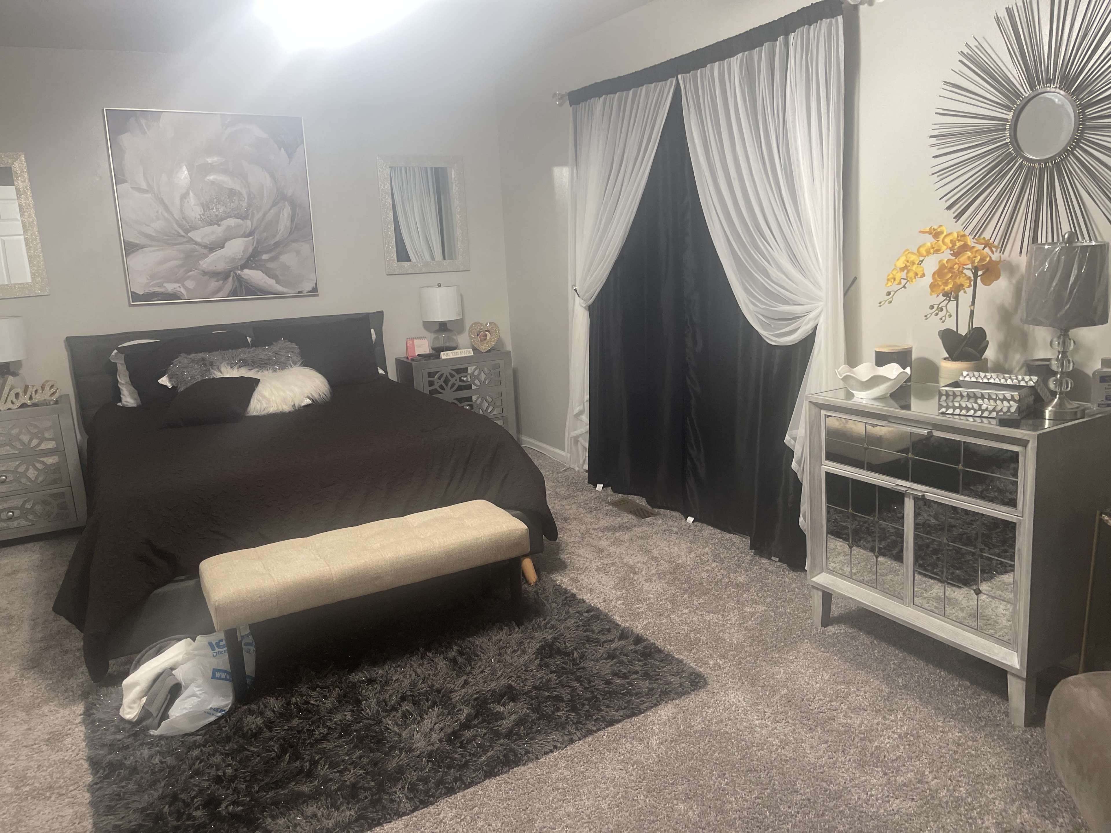 The image shows a bedroom featuring a dark bedspread, a bench at the foot of the bed, two lamps on nightstands, and a dresser with decorative items and a mirror.