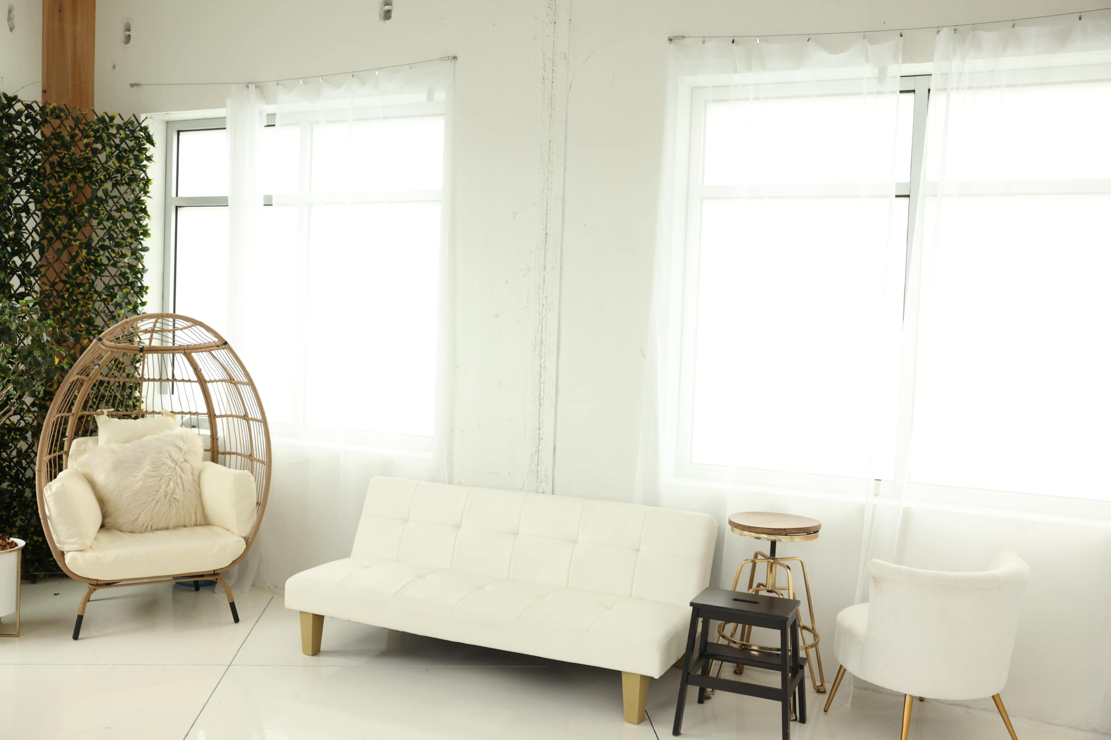The image shows a bright room with large windows, featuring a white sofa, a round chair, and a small wooden table.