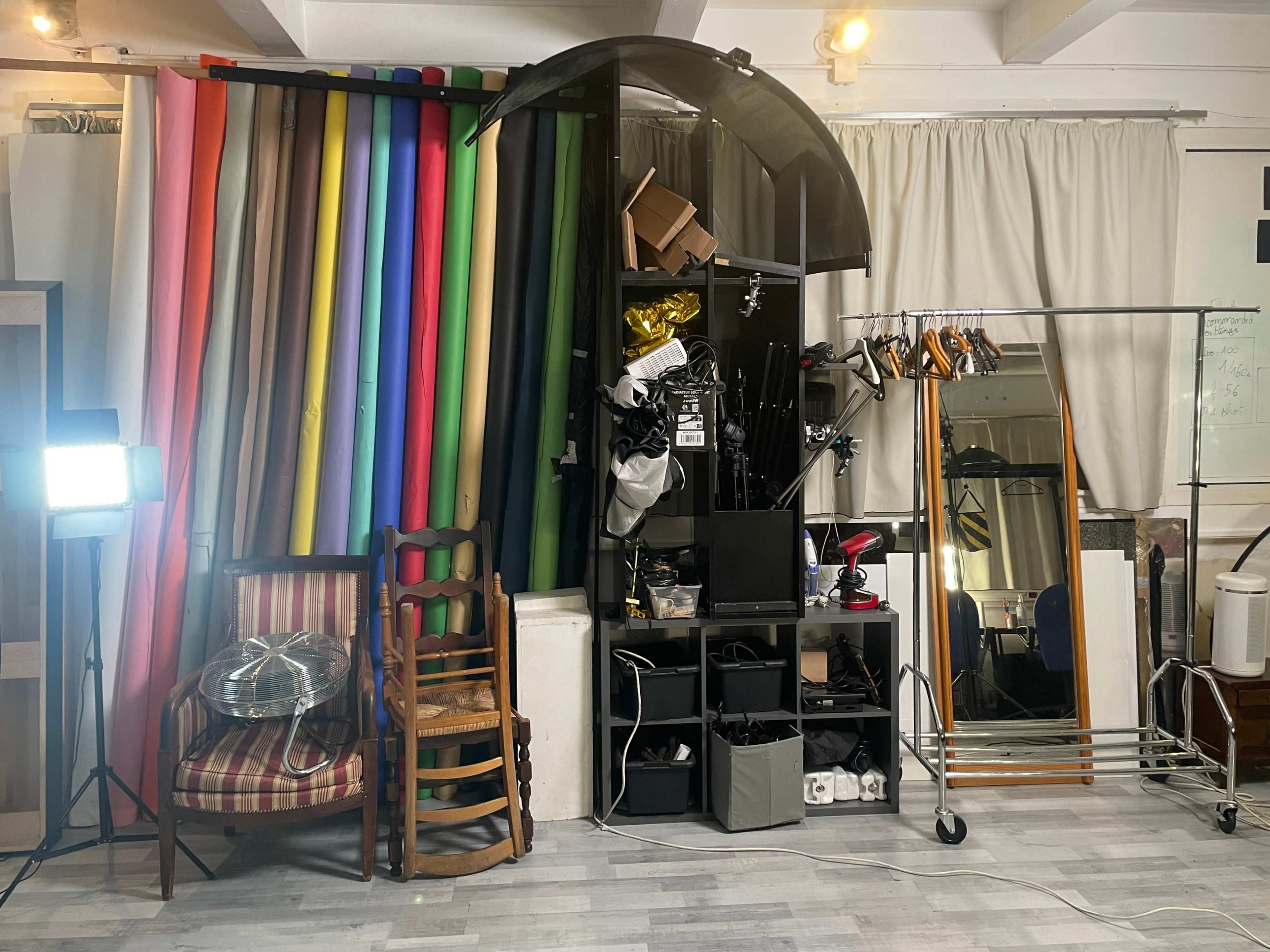 A cluttered studio space with a variety of colorful backdrop rolls, storage shelves filled with equipment, a mirror, and a chair on one side.