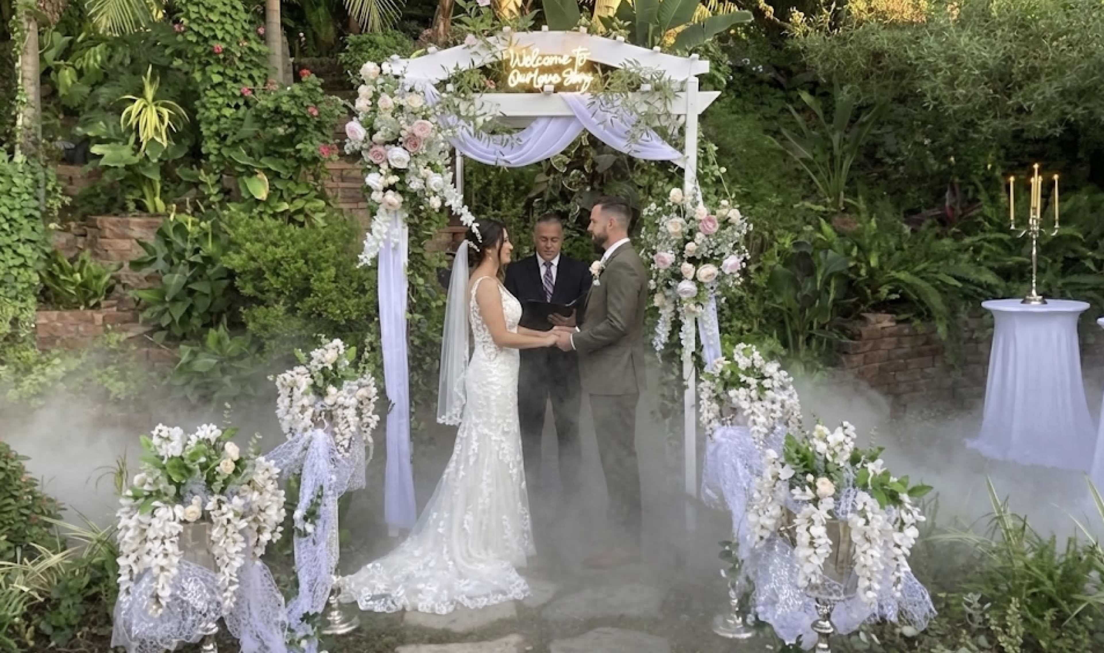 A couple stands under a floral wedding arch, exchanging vows while a celebrant officiates in a garden setting with fog on the ground.