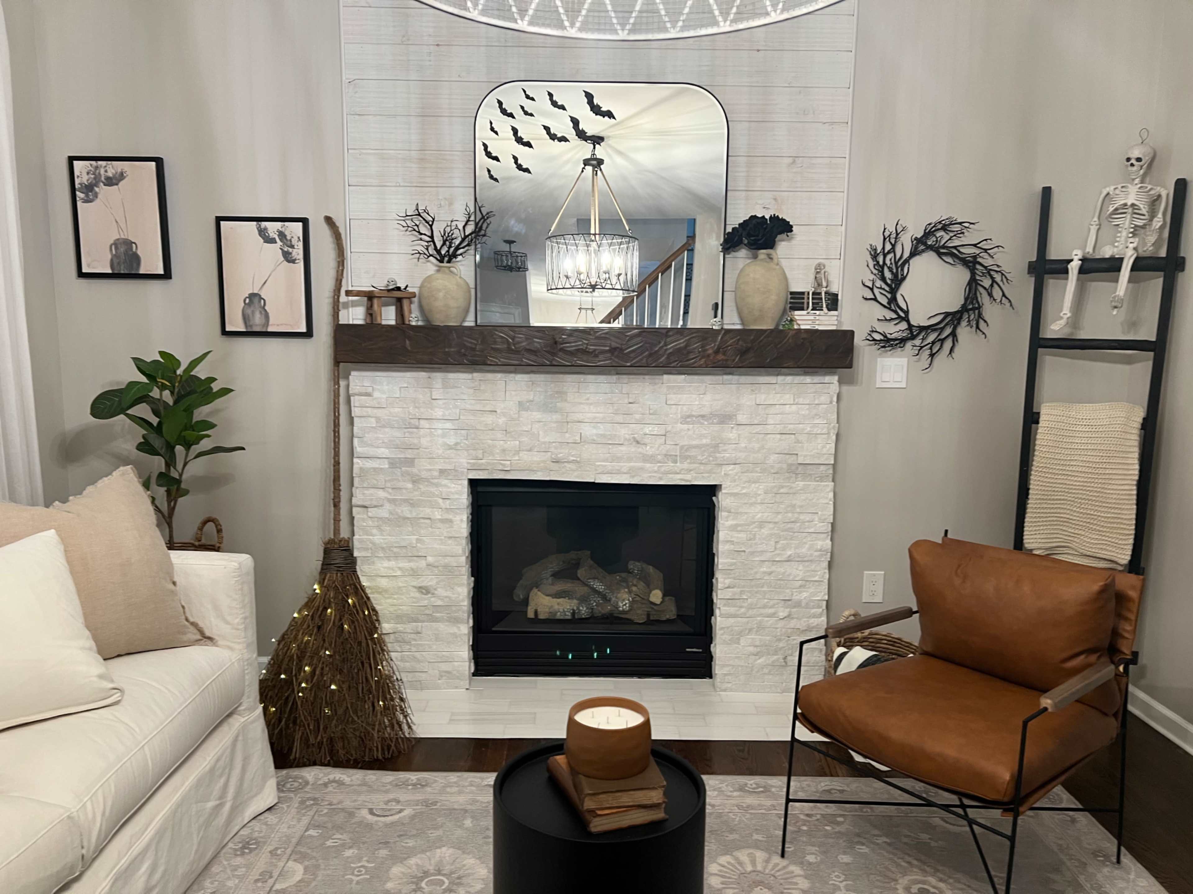 A cozy living room features a stone fireplace, a broom leaning against the mantel, and decorative wall art, with a skeleton and plants adding interest to the space.