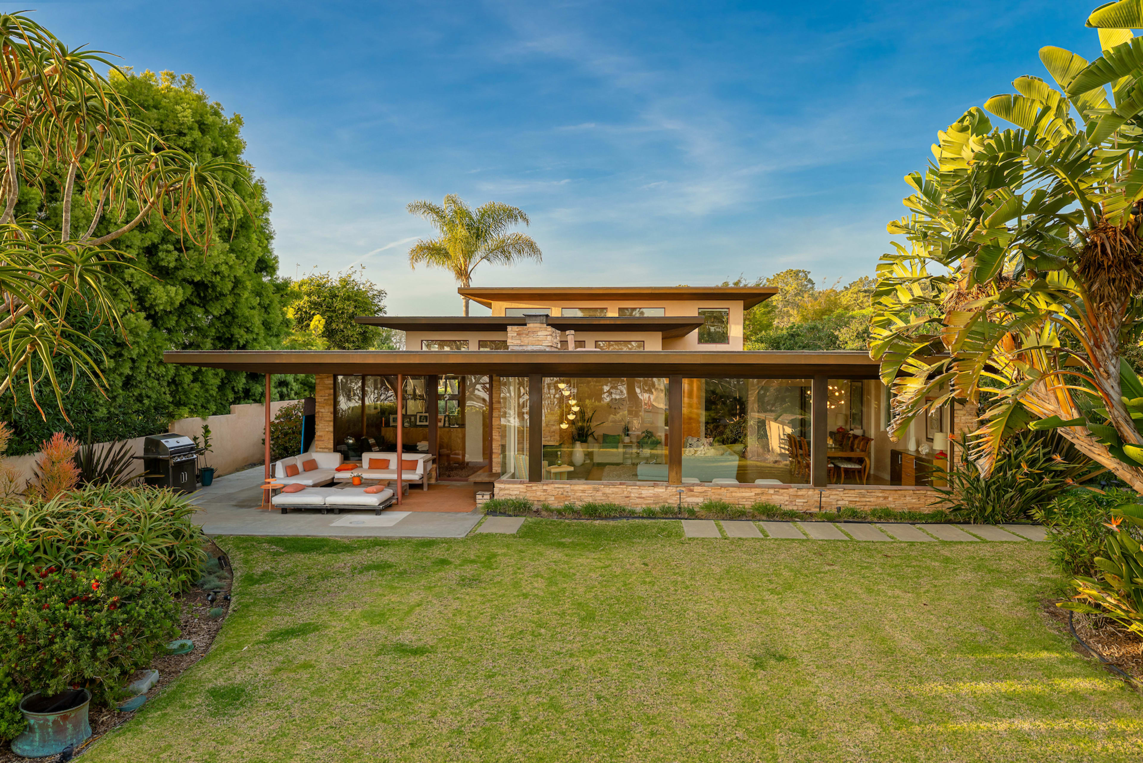 A modern house with large glass windows and a covered patio is surrounded by landscaped greenery.