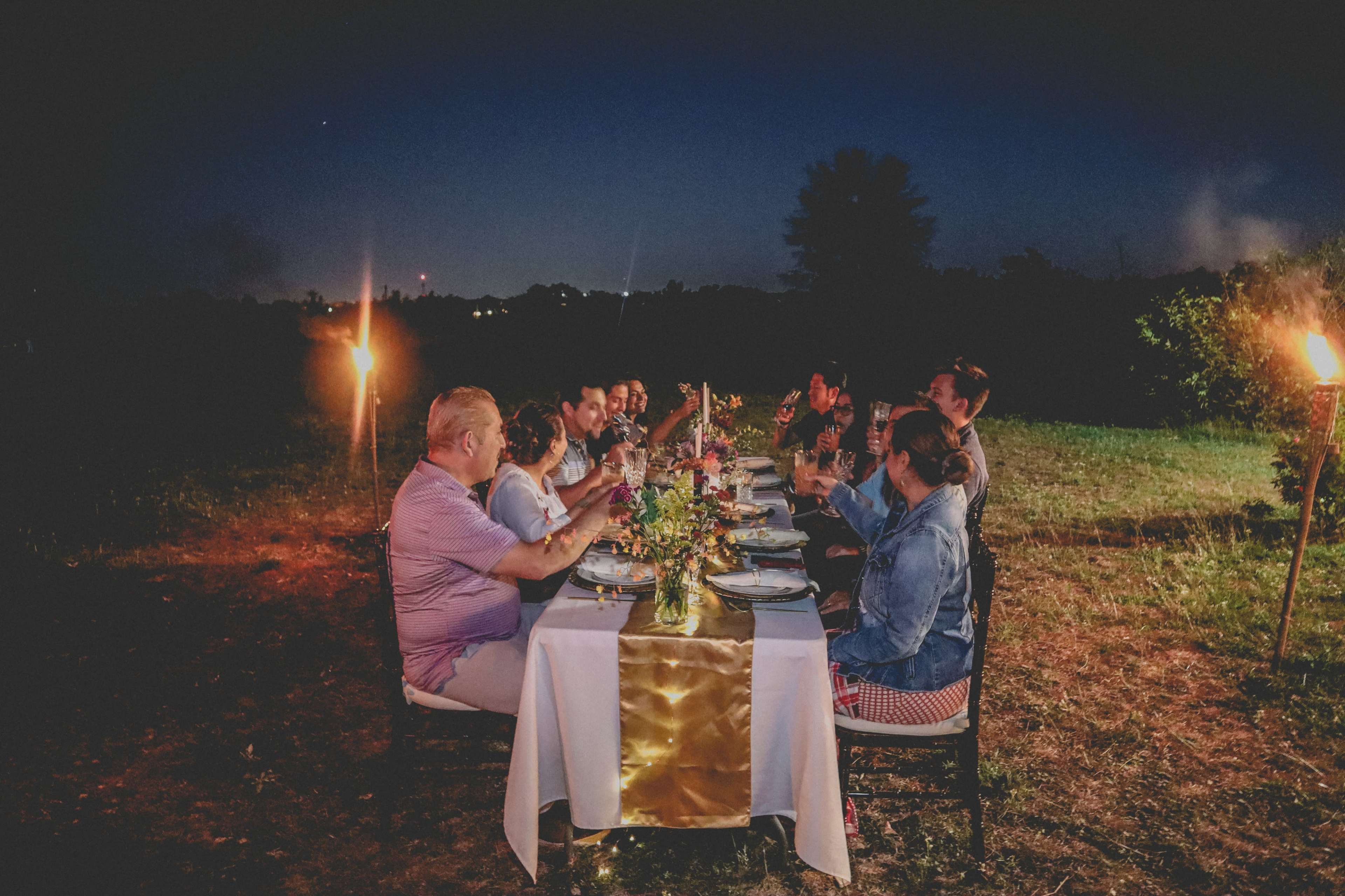 A group of people sits around a long table set for dinner in an outdoor setting at twilight, with tiki torches providing light and a floral centerpiece on the table.