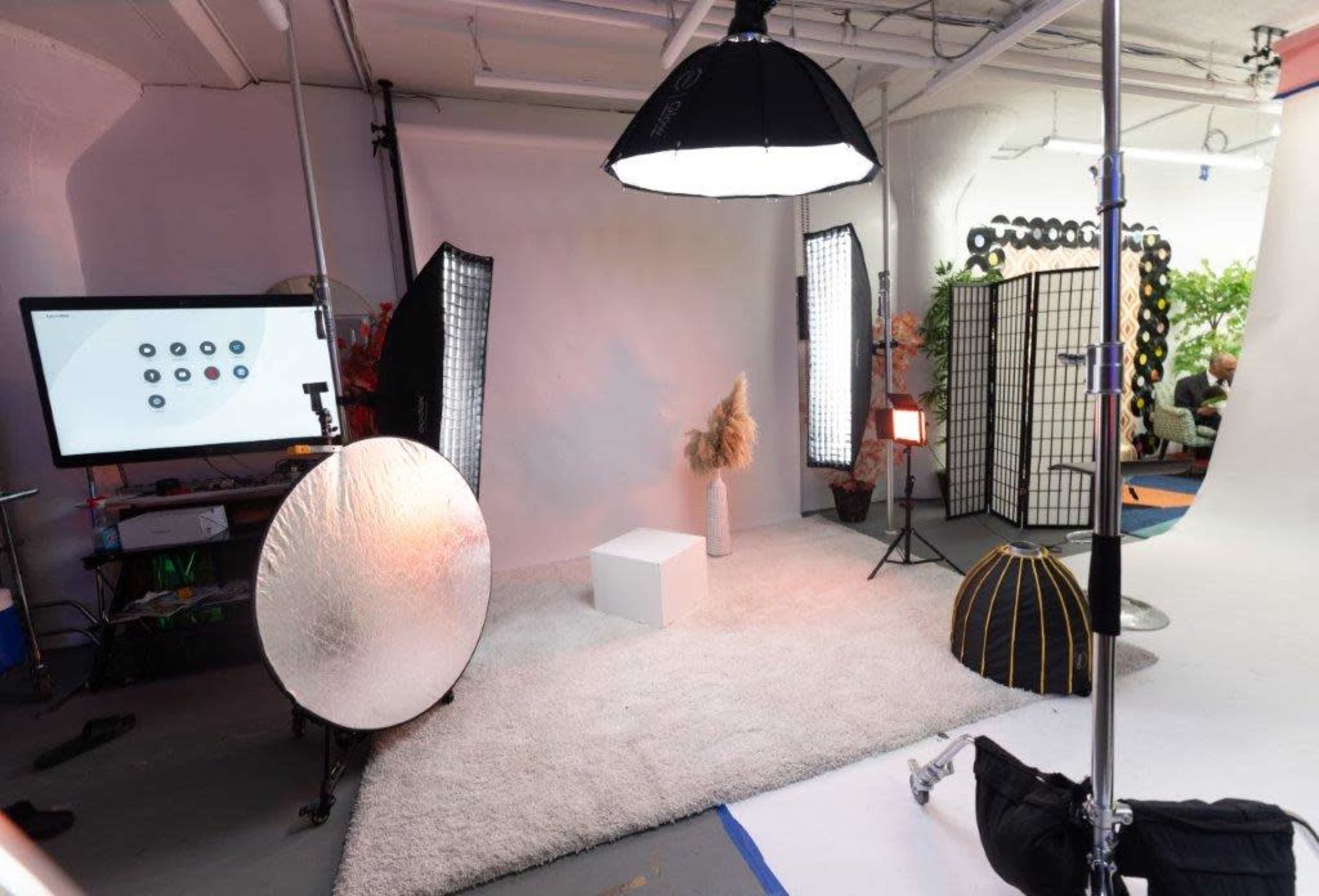 The image shows a photography studio setup with various lighting equipment, a backdrop, and props arranged on a white carpet.