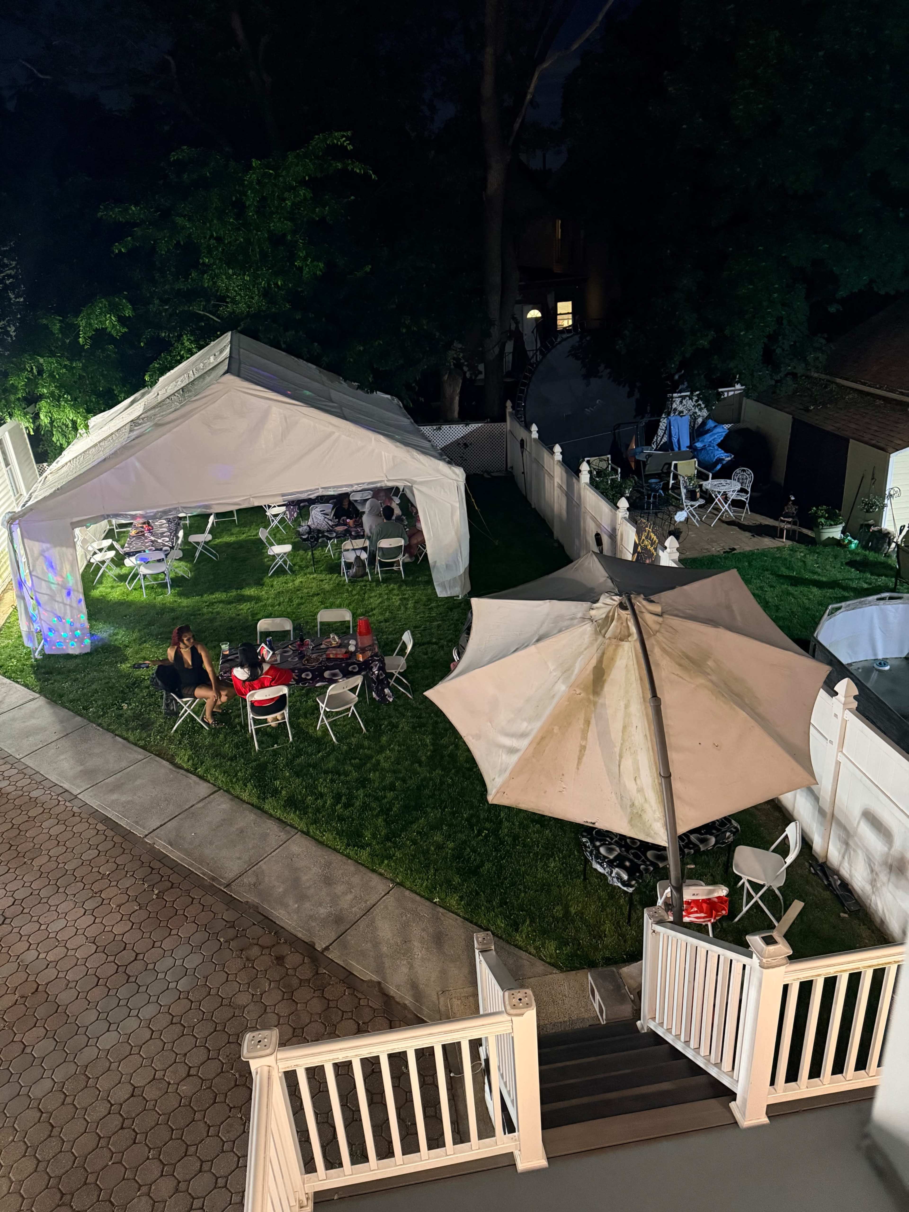 The image shows an outdoor event setup at night, featuring a large white tent, several tables and chairs arranged on grass, and various decorations illuminated by soft lighting.