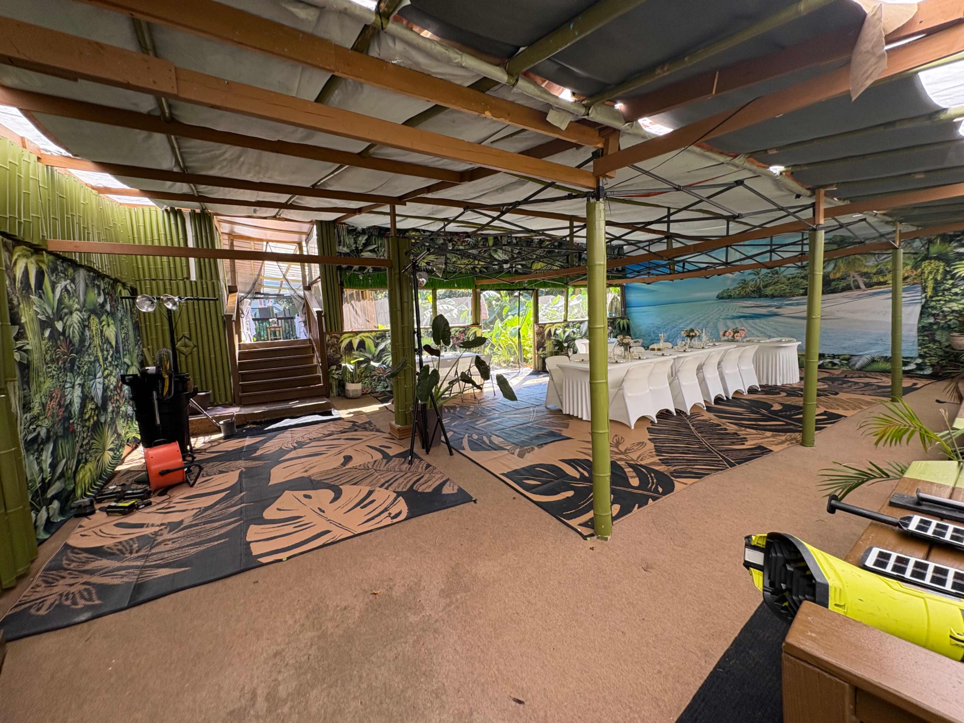 The image shows an indoor event space decorated with tropical murals, featuring tables set for a gathering and a staircase in the background.