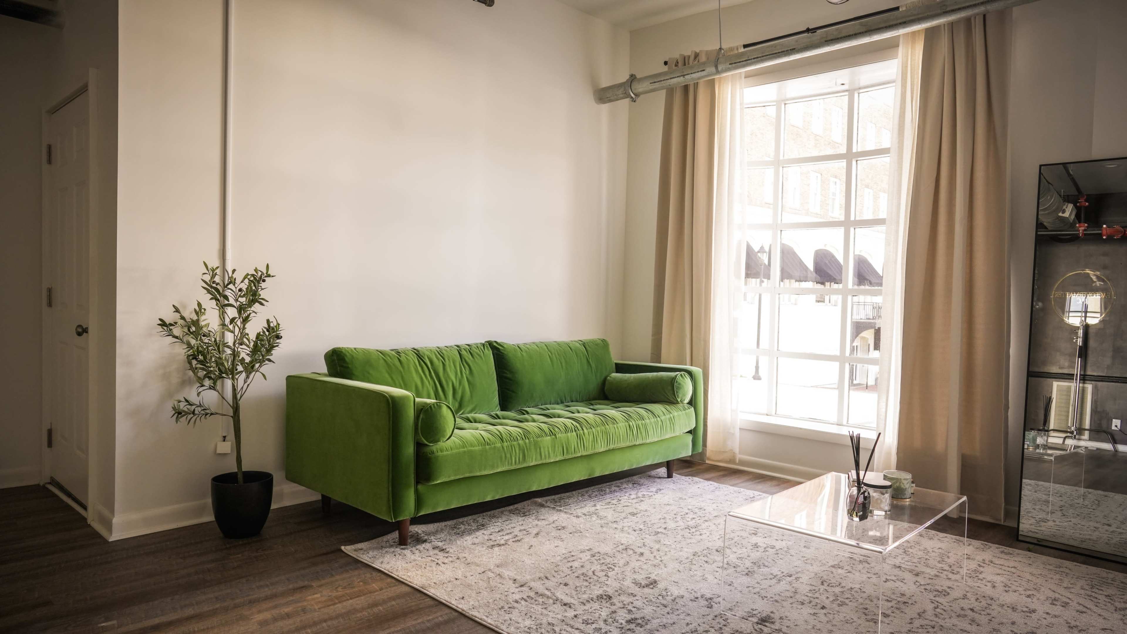 A modern interior with a green sofa, a clear glass coffee table, a potted plant, and large windows with light curtains.