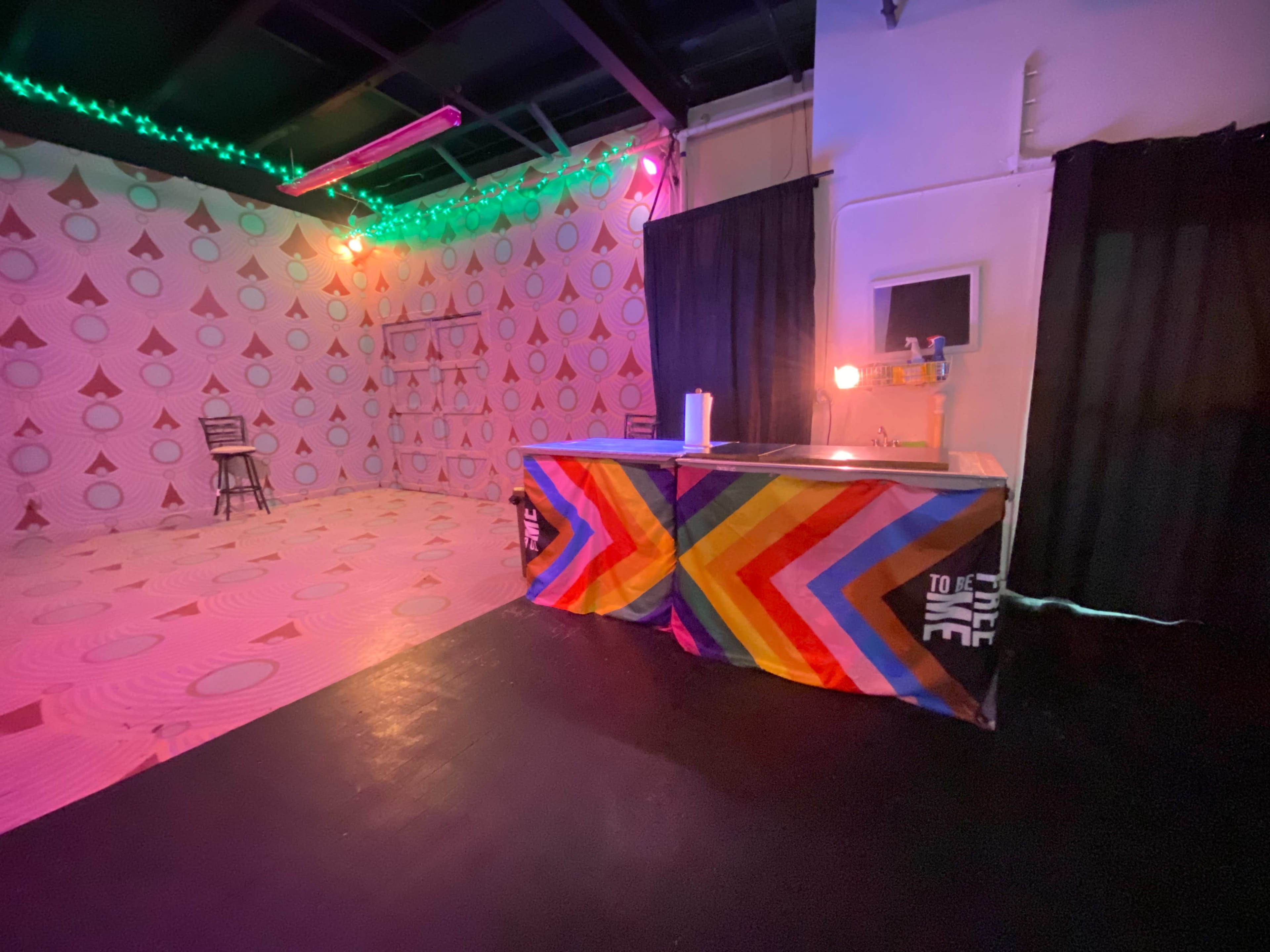 An interior space with brightly patterned walls, a bar with a colorful striped tablecloth, and dim lighting enhanced by green and purple string lights.