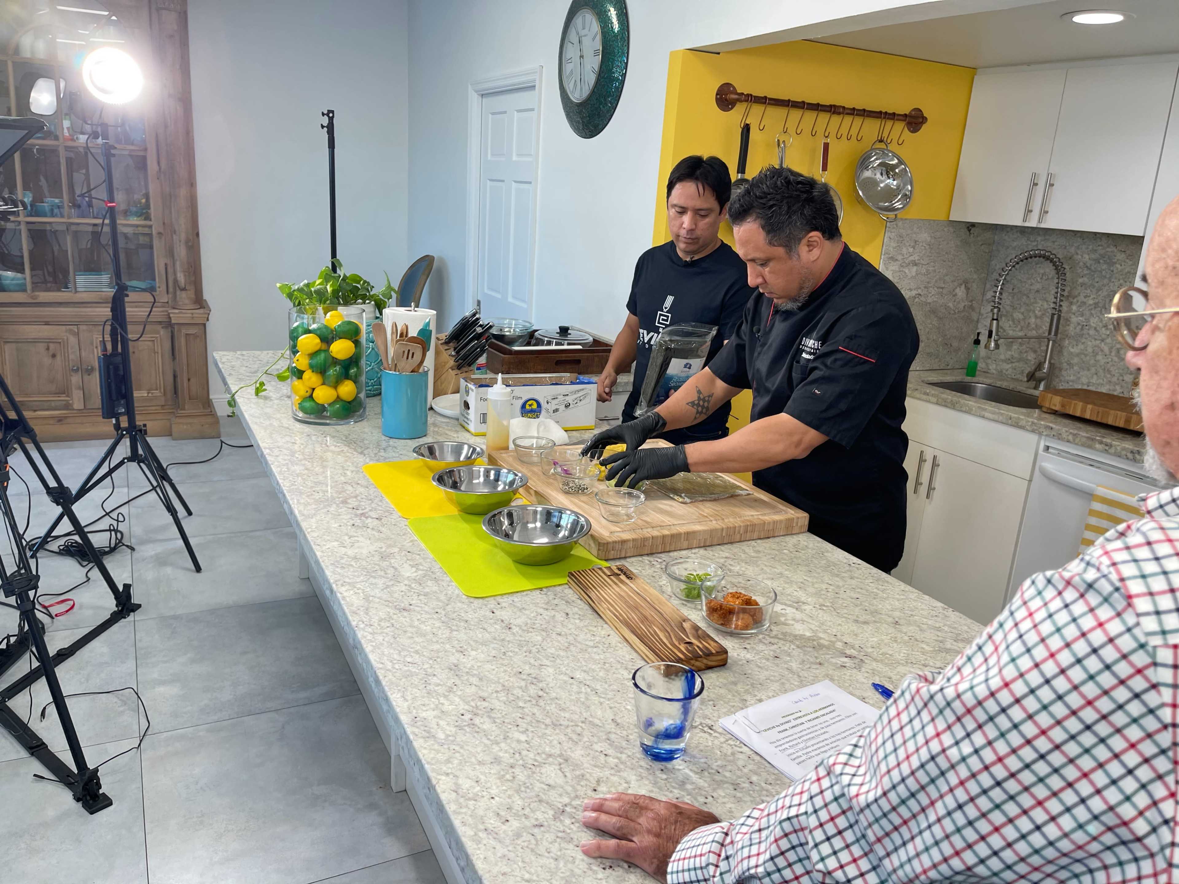 A chef prepares ingredients on a kitchen counter for a cooking demonstration while two observers watch and recording equipment is set up nearby.