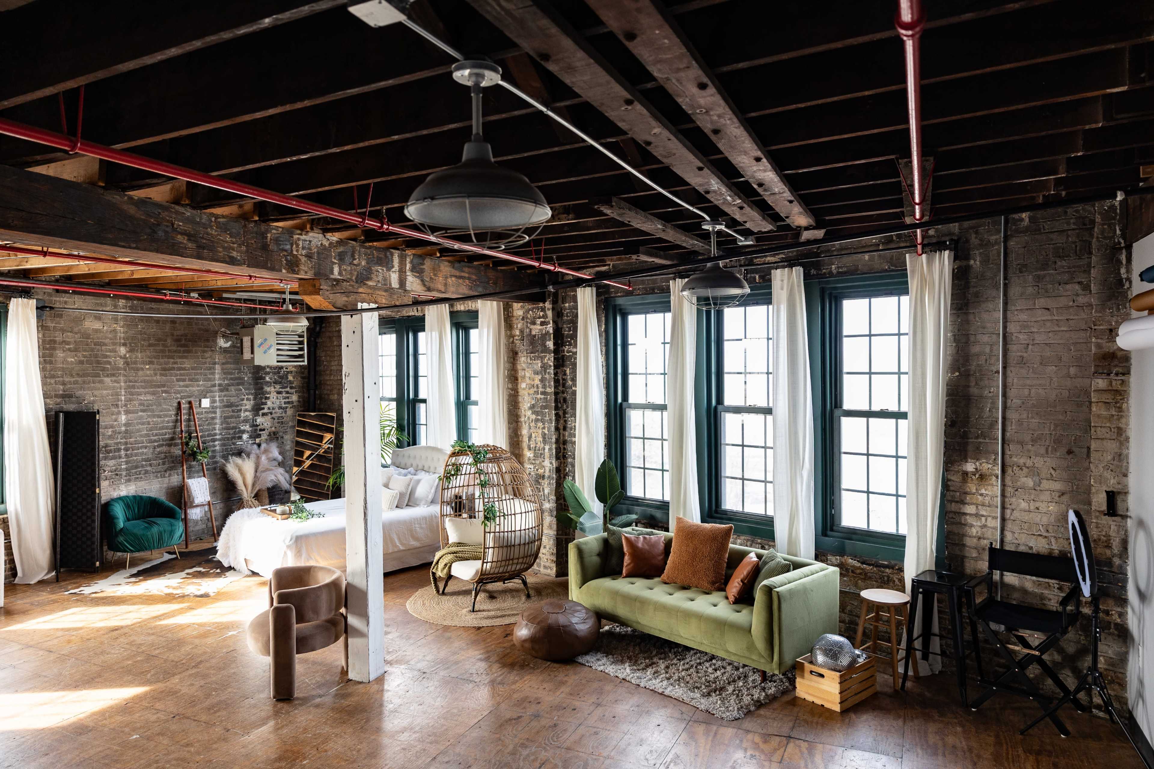 The scene depicts a spacious, industrial-style living area with exposed beams, large windows, and a mix of furniture including a green couch, a bed, and a wicker chair.