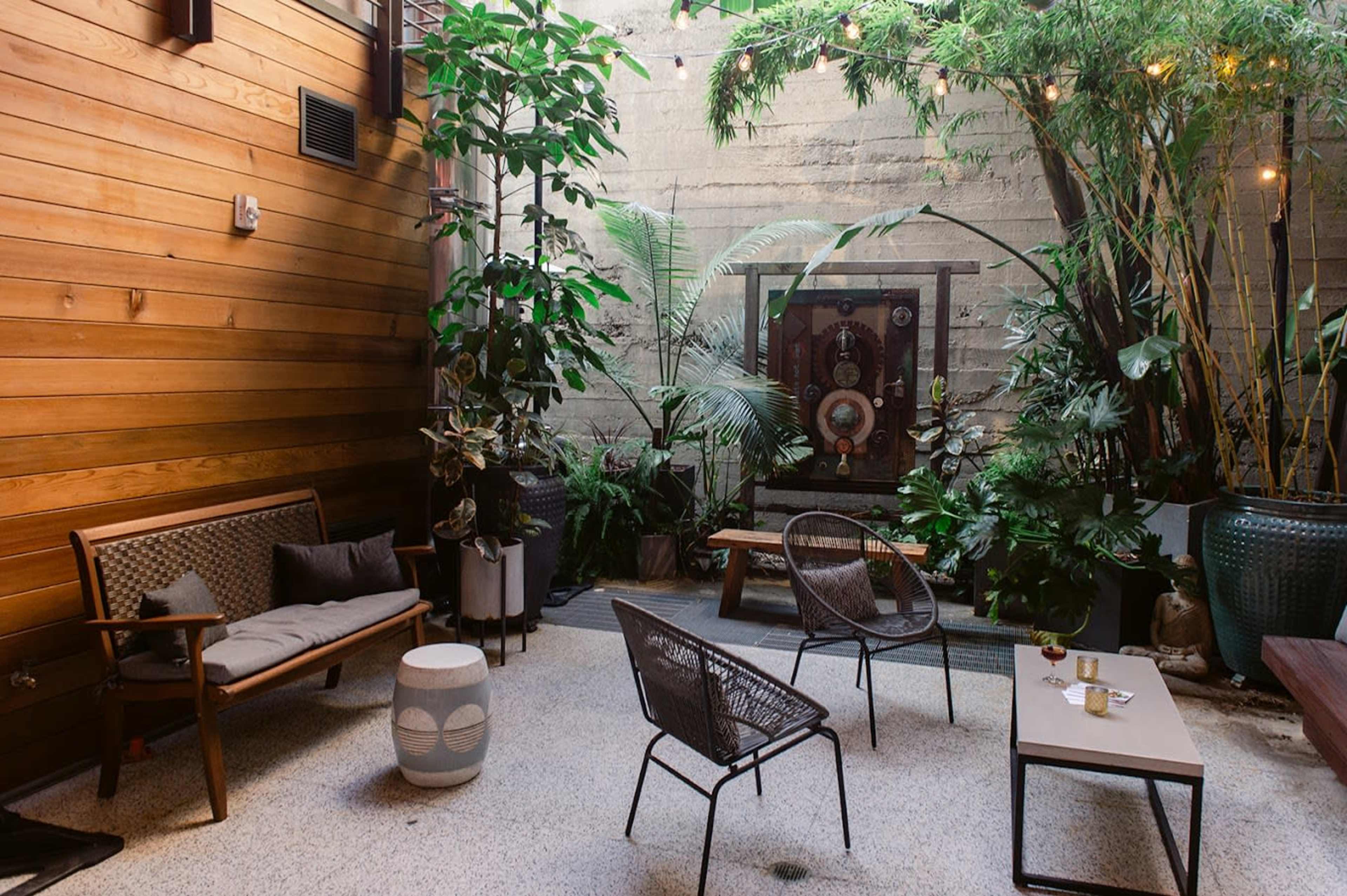 The image depicts a small courtyard featuring wooden walls, various plants, and outdoor seating arrangements.