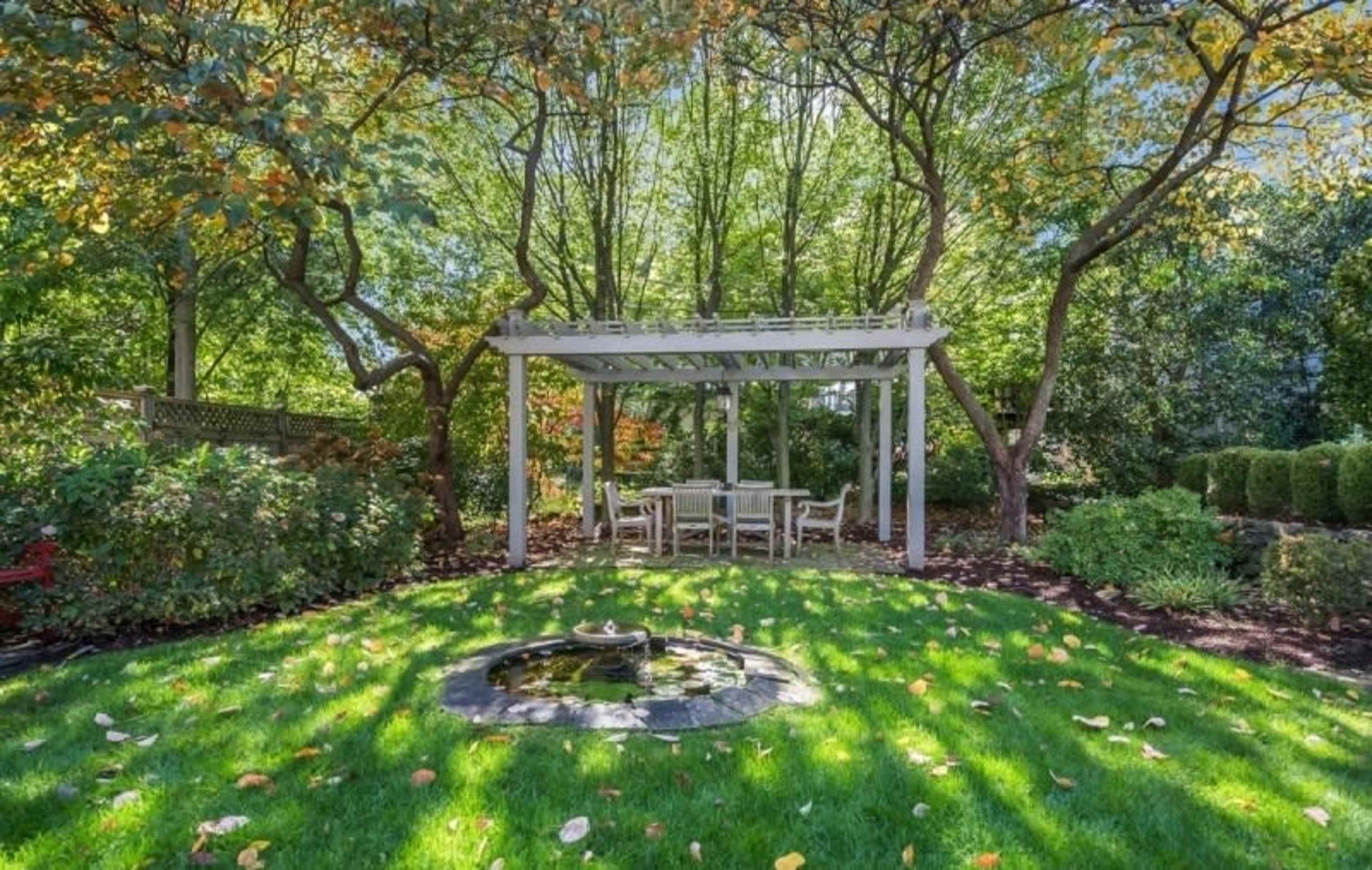 A pergola with a dining set is situated on a grassy area surrounded by trees and garden foliage, with a small fountain nearby.
