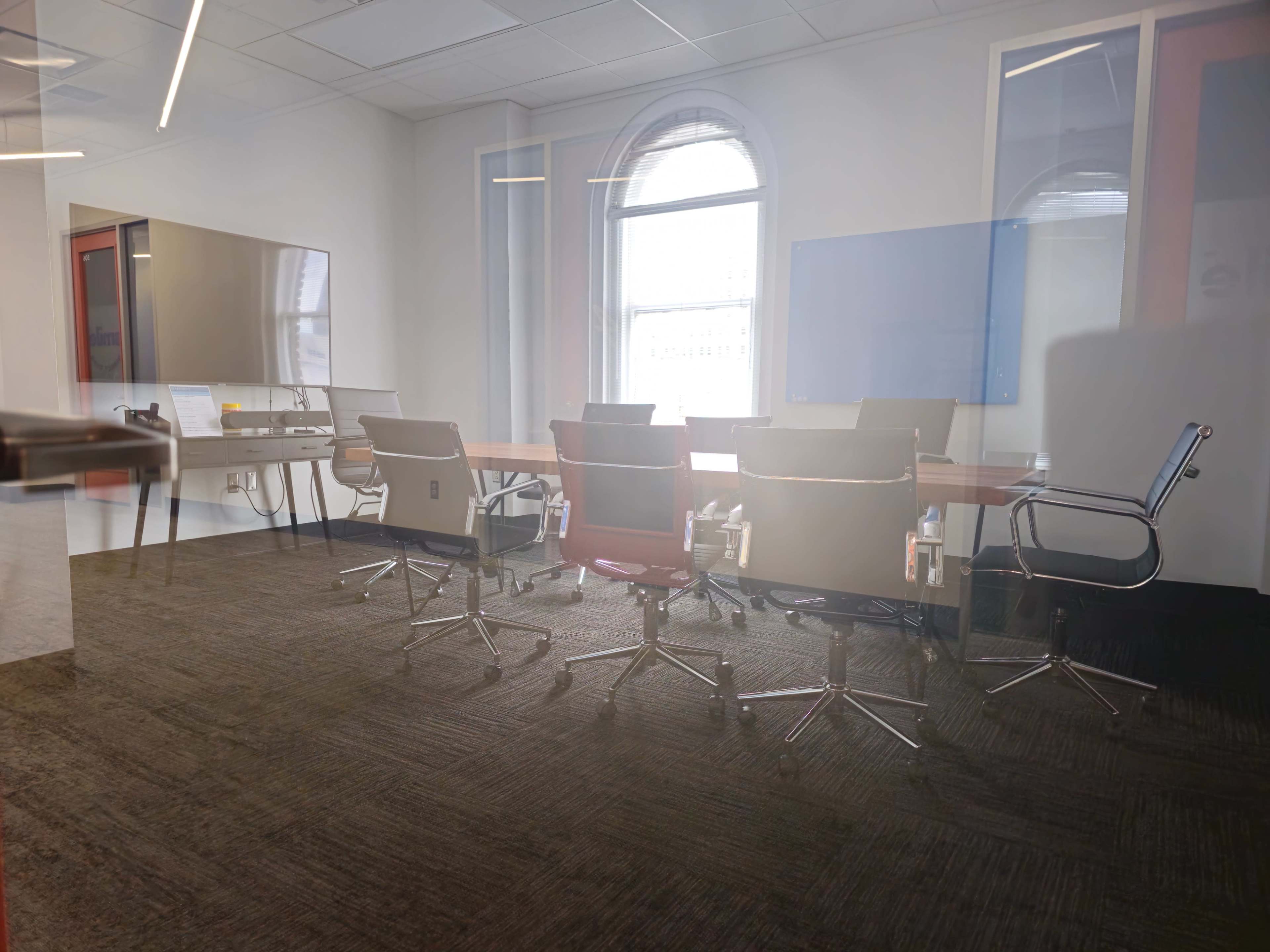 A modern conference room features a long wooden table surrounded by several rolling chairs and a large window providing natural light.